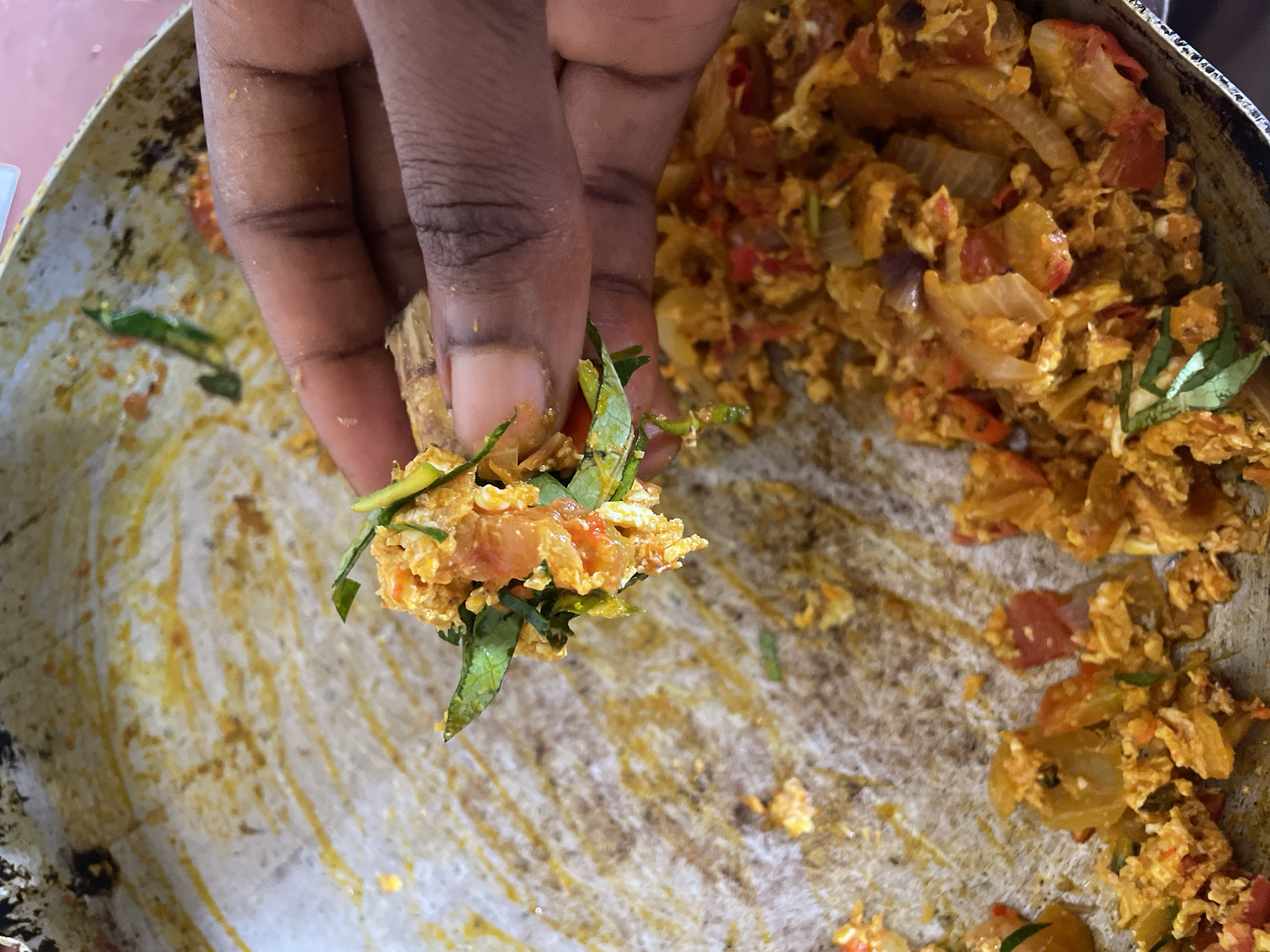A close-up shot reveals a hand lifting a portion of cooked food from a worn, circular pan. The food appears to be a mixture of scrambled eggs, diced tomatoes, and onions, garnished with green leaves. The pan has a light-colored, scratched surface, and remnants of the food are scattered around it. The setting seems to be in a domestic environment. The image was taken in Onitsha, Nigeria.