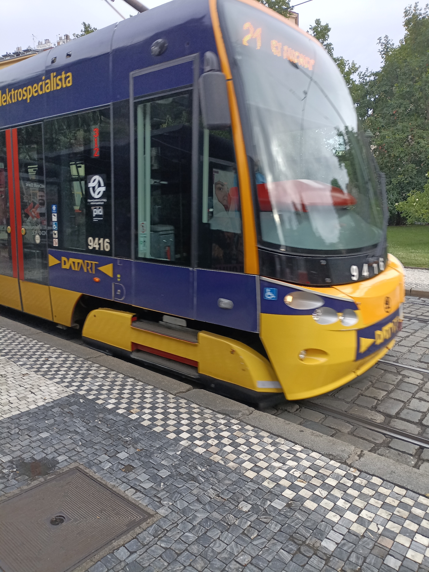 This is a close-up, eye-level, medium shot of a blue and yellow tram moving along a cobblestone street with a black and white checkered sidewalk in Prague, Czechia. The tram has the number "21" displayed in orange LEDs on the front windshield, and the text "ELEKTROSPECIALISTA" is printed in yellow on the blue side of the tram. The tram also displays the number "9416" and the logo "DATART". The front of the tram is yellow and features two headlights and a blue square with a white wheelchair symbol. The cobblestone street is wet, and there is a square metal manhole cover in the foreground. The background is blurred and shows green trees and foliage.