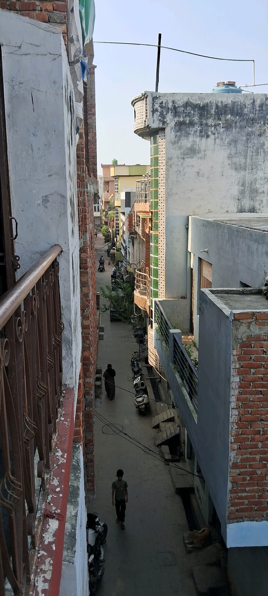 The image depicts a narrow street in Meerut, India, viewed from an elevated position, possibly a balcony or window. The street is lined with multi-story buildings constructed with a mix of brick and concrete, exhibiting various states of repair and architectural styles. On the left, a building features a white-washed exterior with a weathered red-brown railing in the foreground. On the right, a contrasting building showcases a light gray concrete facade with exposed red brickwork at the base, and a balcony with a dark railing.
The street itself is paved and shows signs of use. Several motorcycles and scooters are parked along the sides, and one person is seen riding a motorcycle further down the street. Two individuals are walking in the street: one woman in a dark garment walking away from the viewer, and a young boy in a light olive green t-shirt and dark pants walking towards the viewer. A brown dog is lying down on the side of the street near the right-hand building. The sky is visible at the top, appearing clear and bright. Electrical wires are strung across the street. The overall impression is one of a densely populated urban environment with a mix of residential and commercial structures.