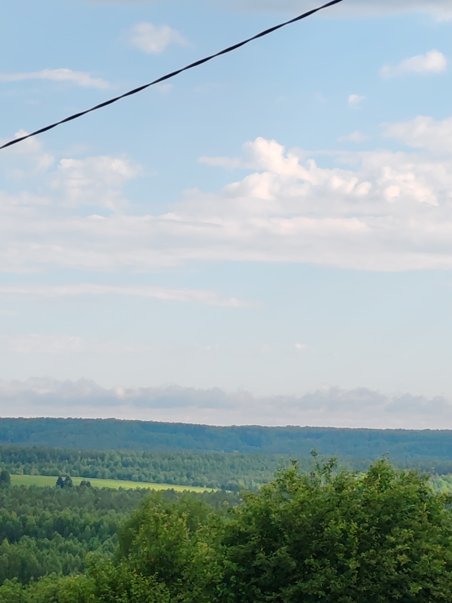 The image shows a vast, undulating landscape under a bright, partly cloudy sky. In the foreground, a dense canopy of green trees fills the bottom portion of the frame. Beyond this, the land slopes downwards and then rises again, covered by a thick, green forest that stretches to the horizon. Interspersed within the forest are patches of bright green, suggesting clearings or fields. A faint, dark, diagonal line, likely a power line, cuts across the upper portion of the sky. The overall impression is of a rural or natural environment. While the image does not explicitly provide the location in Chukhloma, Russia, it depicts a typical landscape found in such a region, characterized by extensive forests and rolling terrain.