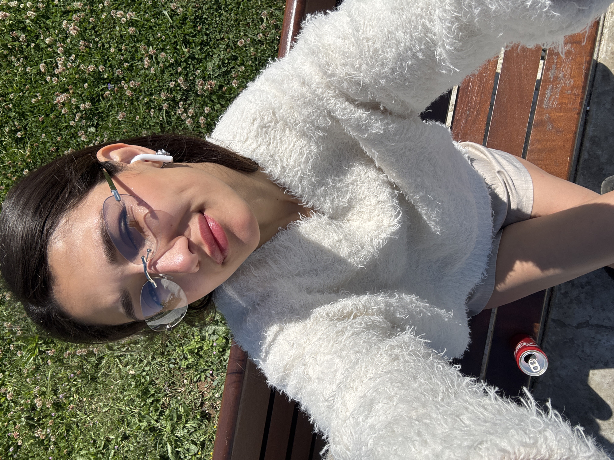 A young woman is sitting on a wooden bench outdoors. She is wearing a white, fluffy sweater, beige shorts, and unique glasses with clear lenses and silver frames. She has dark hair and is wearing a white earbud in her left ear. An open can of Coca-Cola is on the bench near her. The bench is located near a grassy area with clover. It appears she is taking a selfie. While the request mentions Šušanj, Montenegro, this cannot be confirmed solely from the image.