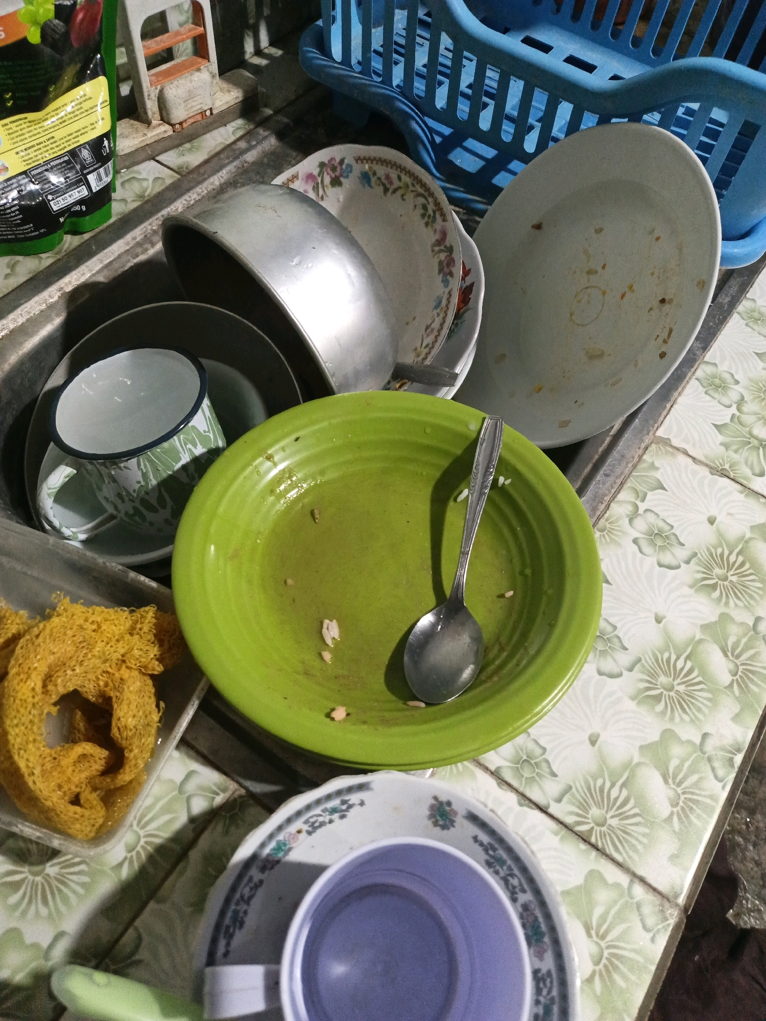The image depicts a kitchen sink area with various dirty dishes and kitchenware, set in the city of Keleyan, Indonesia. The sink is filled with a few plates, bowls, and a mug, some of which have food residue. A green plate with a spoon is prominently displayed in the foreground. To the left, a yellow sponge is visible. In the background, a blue plastic dish rack is partially seen, containing more dishes. The surrounding countertop is tiled with a floral pattern in green and white. There are no people visible in the image, and the scene suggests a domestic setting where cleaning is yet to be done.