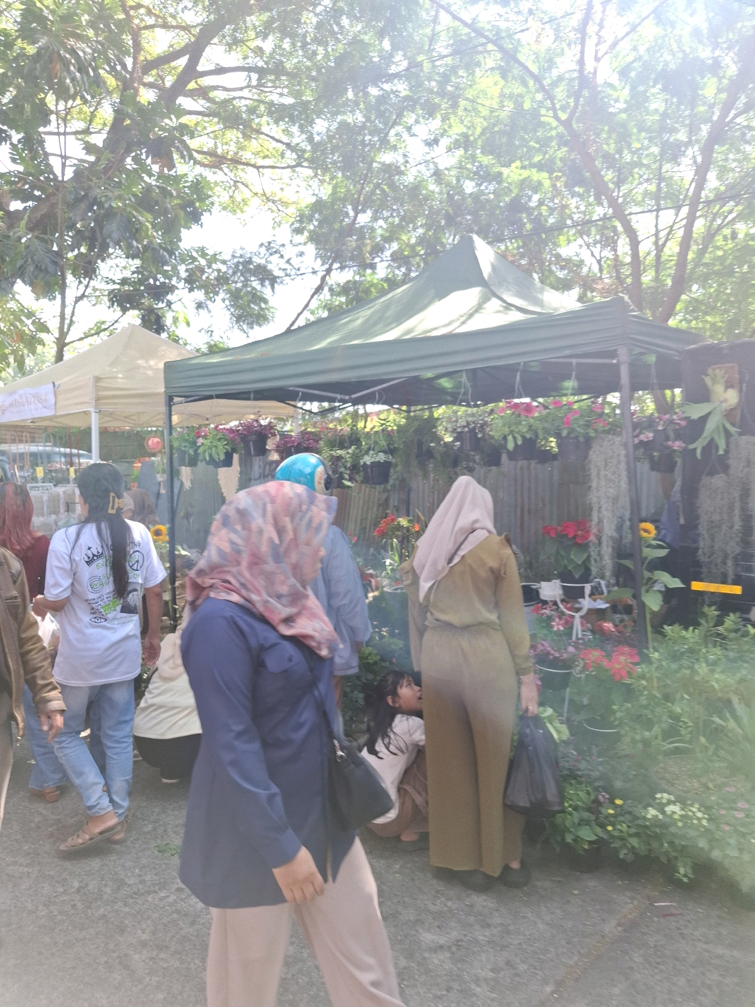 In Karangtengah, Indonesia, an outdoor marketplace is captured in daylight under a canopy of trees. A green tent, erected for a vendor, displays an assortment of potted plants, including red flowers and hanging greenery. To the left, a lighter colored tent is visible, suggesting multiple vendors or a larger market setup. Several individuals are present. In the foreground, a person in a navy blue top and light brown pants walks away from the camera, their face obscured by a patterned headscarf. To their left, a person in a light blue shirt and jeans, with dark hair pulled back, looks towards the tents. Further left, another individual in a brown jacket and jeans is partially visible. In the middle ground, a child wearing a light-colored shirt crouches down. Another person in a long, light brown outfit and a beige headscarf stands near the child, holding a black plastic bag. The ground is a paved surface, appearing to be concrete or asphalt. The scene is illuminated by bright sunlight, creating a warm atmosphere.