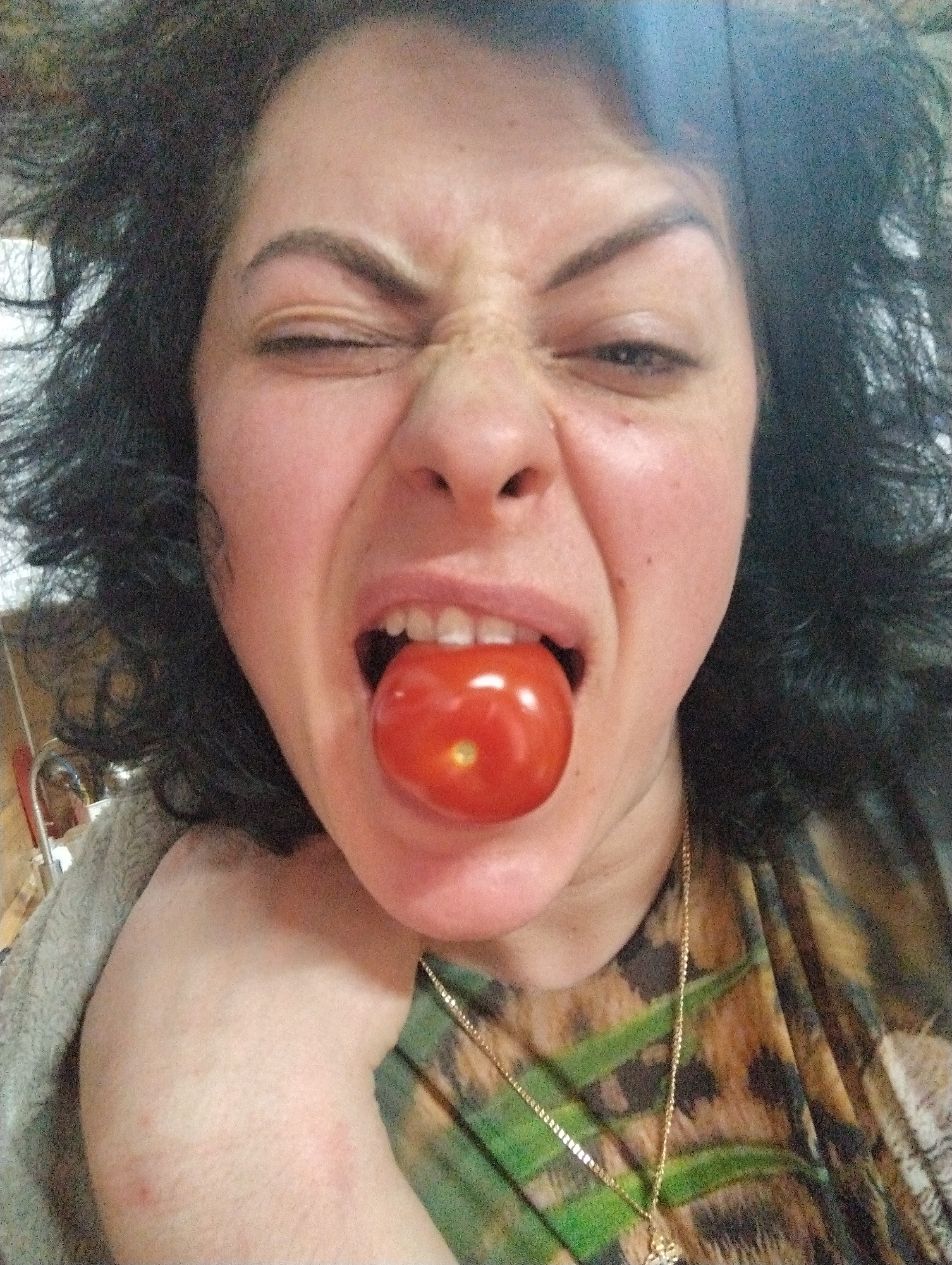 A woman is positioned in the foreground with her head and shoulders visible. She is making a distorted facial expression, with her eyes squinted, nose wrinkled, and mouth open with a tomato inside. She has dark curly hair and is wearing a printed shirt and a gold necklace. Part of her arm is visible on the left side of the frame, covered with a gray shawl. The background is blurry and appears to be an interior space with furniture visible on the left side of the frame. It is not possible to verify this is in Grishanki, Russia.