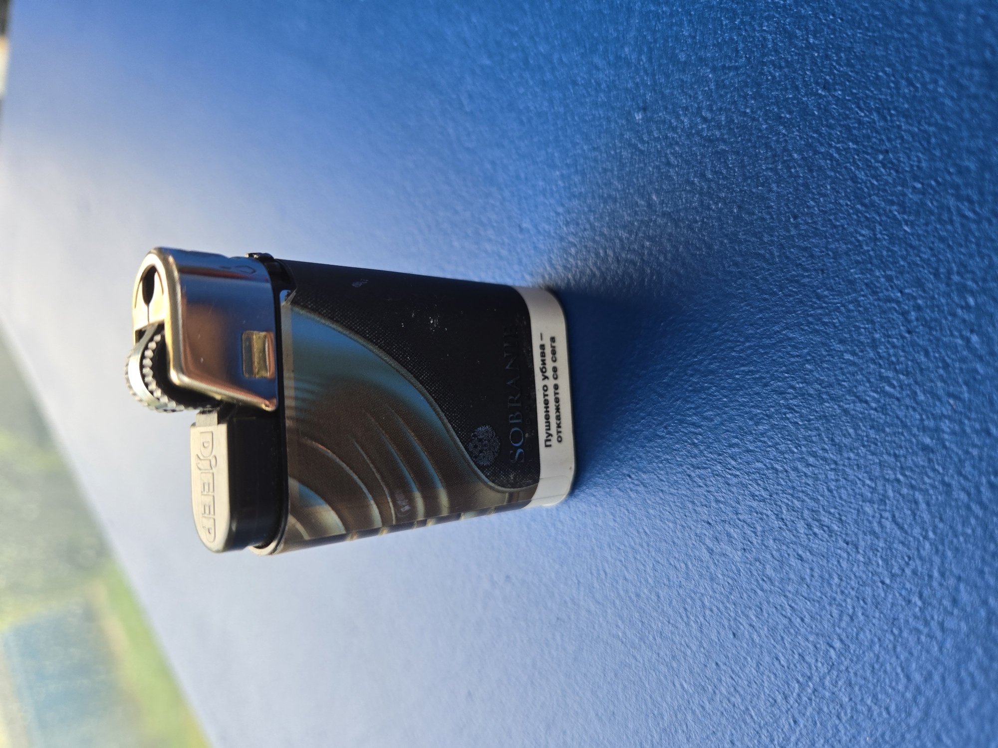 A close-up image displays a lighter resting horizontally on a vibrant blue, textured surface. The lighter features a metallic, chrome-plated top mechanism with a prominent striking wheel. The main body of the lighter is black, embellished with an artistic design of swirling lines in shades of gold, brown, and dark grey. The brand name "Pjeep" is visibly embossed on the chrome component of the lighter.
A white label on the lighter's side includes partially visible text: "SOBRAKOL" (likely part of "SOBRANIE"). Below this, in Cyrillic script, is the phrase "Пушенето убива - откажете се сега", which translates to "Smoking kills - quit now".
The background is a deep blue surface with a distinct, rough, and granular texture, possibly painted concrete or stucco. The scene is brightly illuminated by strong, direct light, suggesting it was captured outdoors during daytime under sunny conditions. This lighting creates sharp shadows and high reflections on the lighter's metallic parts. Reflections in the chrome also show blurry patches of lighter blue and green, indicating an open environment, perhaps reflecting the sky or distant foliage. No activity is taking place; the image is a static portrayal of the lighter.