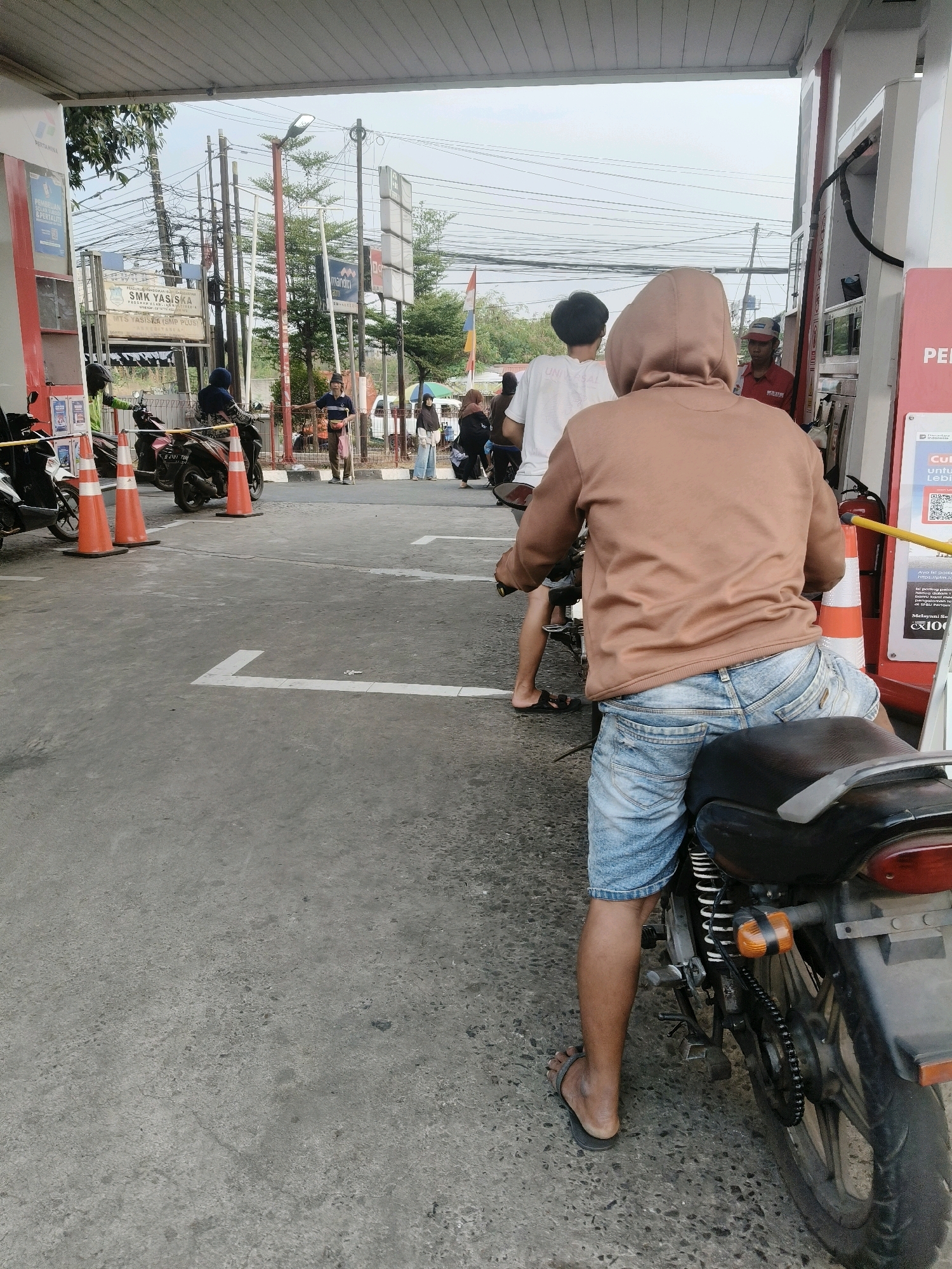 The image depicts an outdoor scene at a Pertamina gas station in South Tangerang, Indonesia. In the foreground, a person wearing a tan hoodie and blue denim shorts sits on a motorcycle, facing away from the camera. The motorcycle is black and has visible suspension and a chain. The ground is paved with asphalt and has white lane markings.
In the middle ground, several other individuals are present, also near motorcycles. Some are queuing, while others appear to be waiting. There are orange traffic cones placed on the ground. Behind them, there are various signs, including one for "SMK YASISKA". A gas pump station is visible on the right side of the image.
The background shows a street with utility poles and electrical wires, trees, and buildings. The sky is overcast. The overall impression is a busy everyday scene at a public service location.