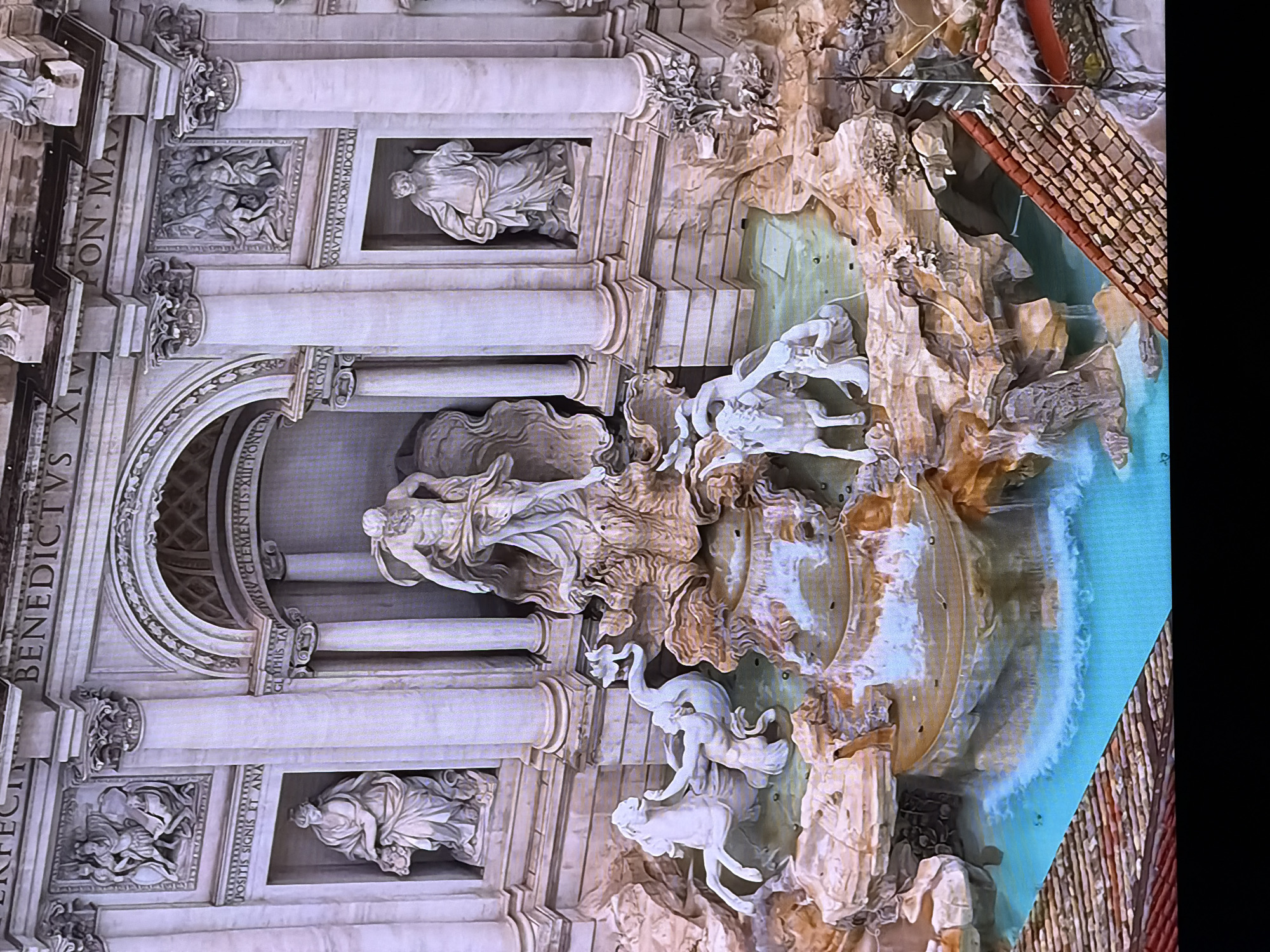 The image shows a close-up view of the Trevi Fountain in Rome, Italy, not Chirchik, Uzbekistan. It is a Baroque-style fountain, the largest Baroque fountain in the city and one of the most famous fountains in the world. The fountain is dominated by a central figure of Oceanus, god of the sea, riding a shell-shaped chariot pulled by two sea-horses. Flanking Oceanus are statues of Abundance and Salubrity. The entire composition is set against the backdrop of the Palazzo Poli, which houses the Aqua Virgo aqueduct. The foreground of the image shows the water cascading down the rocky, sculpted landscape of the fountain, creating a dynamic and dramatic effect. The water is a clear, turquoise blue, with white foam and spray where it crashes against the rocks. The architectural elements of the Palazzo Poli are visible in the background, including columns, niches with statues, and decorative carvings. The image captures the grandeur and artistry of the Trevi Fountain, highlighting its intricate details and the movement of water.