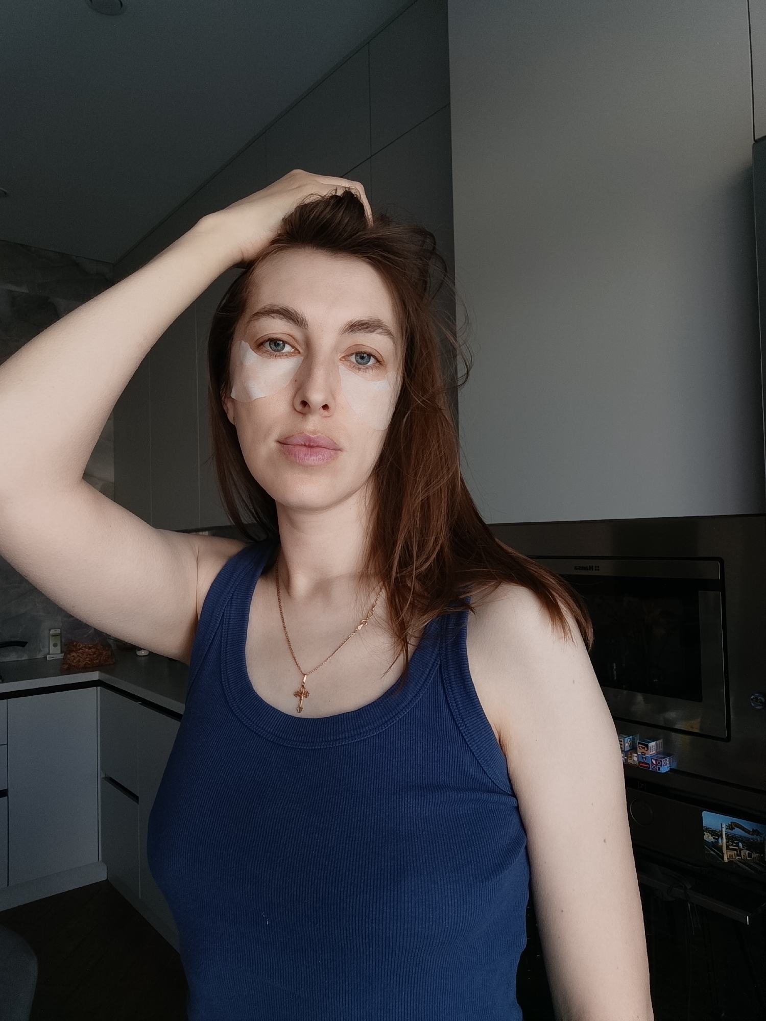 A woman is posing indoors. She is fair-skinned with blue eyes and brown hair. She wears a blue tank top and a gold necklace with a cross. She has adhesive eye patches under both eyes. Her left arm is raised, with her hand touching her hair. A modern kitchen is visible in the background with white cabinets and a stainless steel oven, and there is something on the oven door, possibly magnets. The location is unknown; it is unlikely to be possible to ascertain that the image was taken in Chaikovsky, Russia, from the visual information provided.