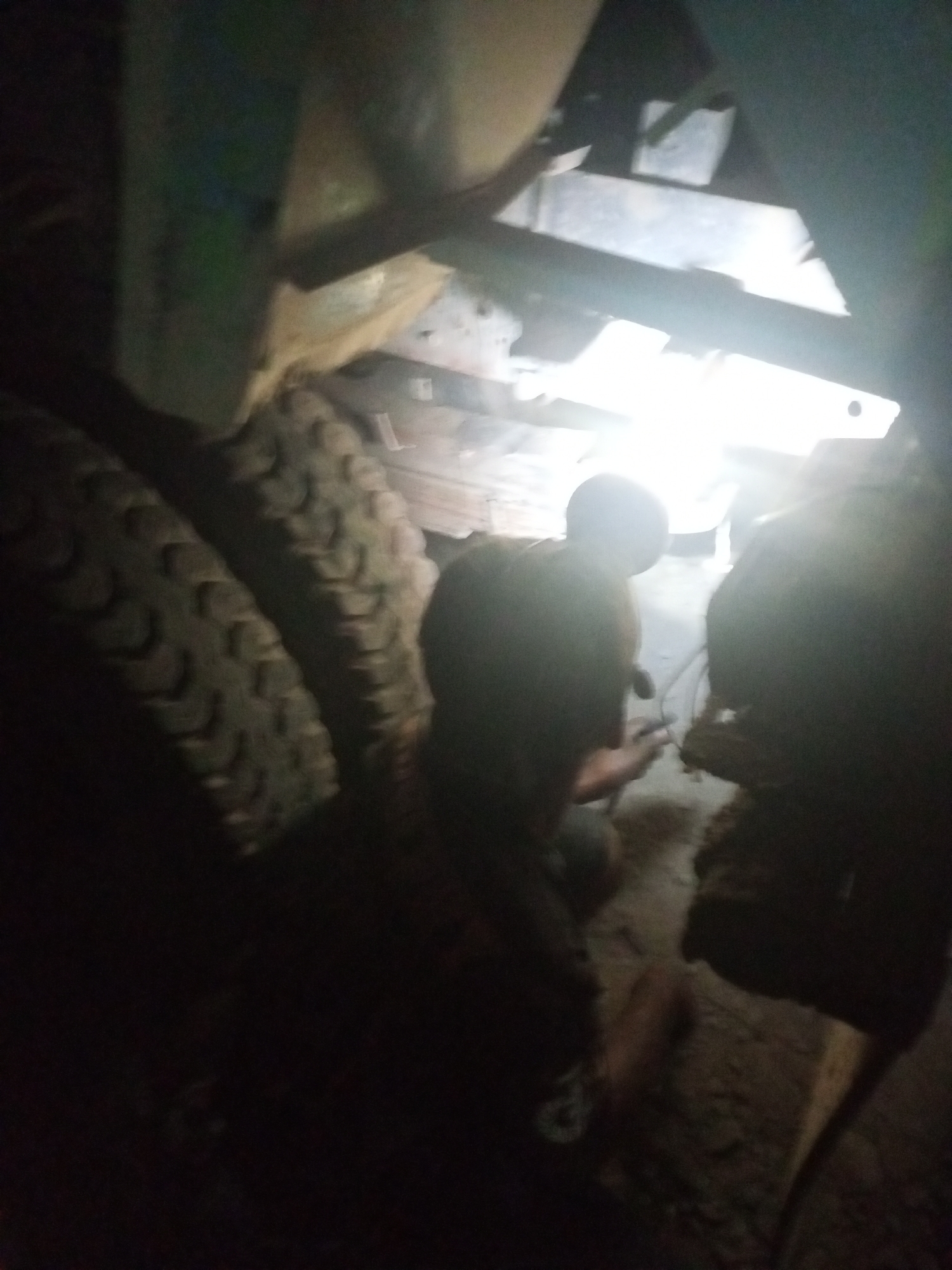 In Keling Panggau, Indonesia, an image captures a dimly lit scene under a large vehicle. Two individuals, silhouetted and partially obscured, are working on the underside of the vehicle. One person appears to be bent over, possibly attending to mechanical components, while the other is partially visible to their right. The ground is uneven and appears to be dirt or gravel. Two large tires are prominently visible in the foreground on the left side of the image. The overall lighting is low, with bright light emanating from the vehicle's undercarriage, creating strong contrasts and obscuring finer details.