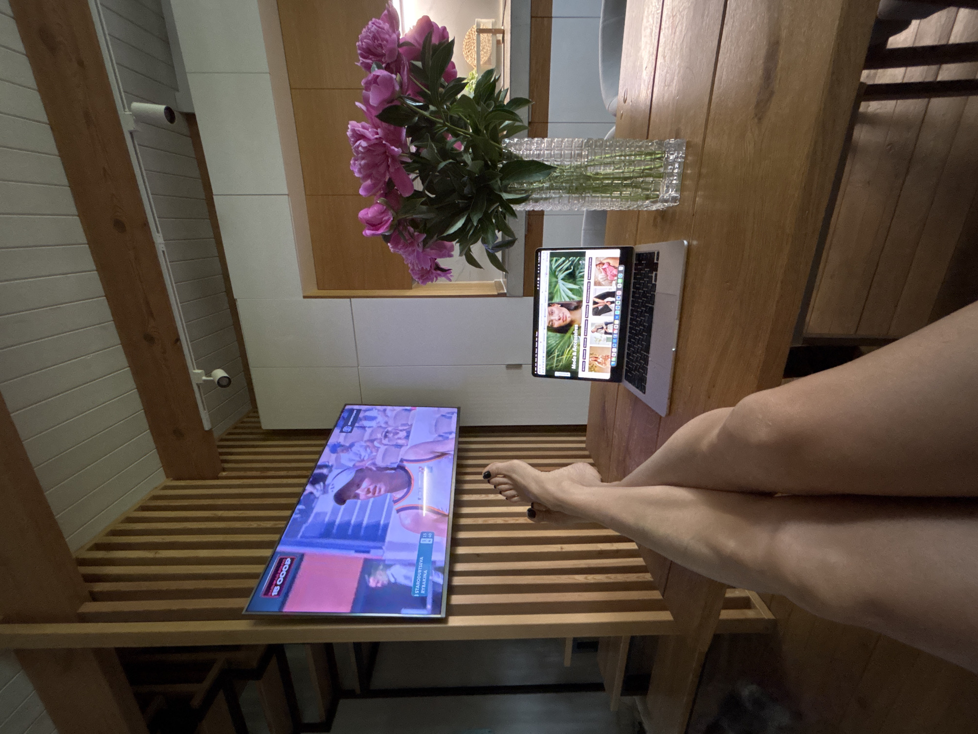 The image shows an indoor scene with a combination of work and leisure elements. A woman's legs are crossed, suggesting relaxation, while a laptop displaying a website is open on a wooden table alongside a vase of pink flowers. A television displaying a sporting event is positioned on a lower wooden surface, suggesting the presence of an ongoing broadcast. The location appears to be a domestic interior, possibly a modern apartment, evidenced by the wooden structure and minimalist décor. The location is not specified as the city of Chaikovsky, Russia, from the scene.