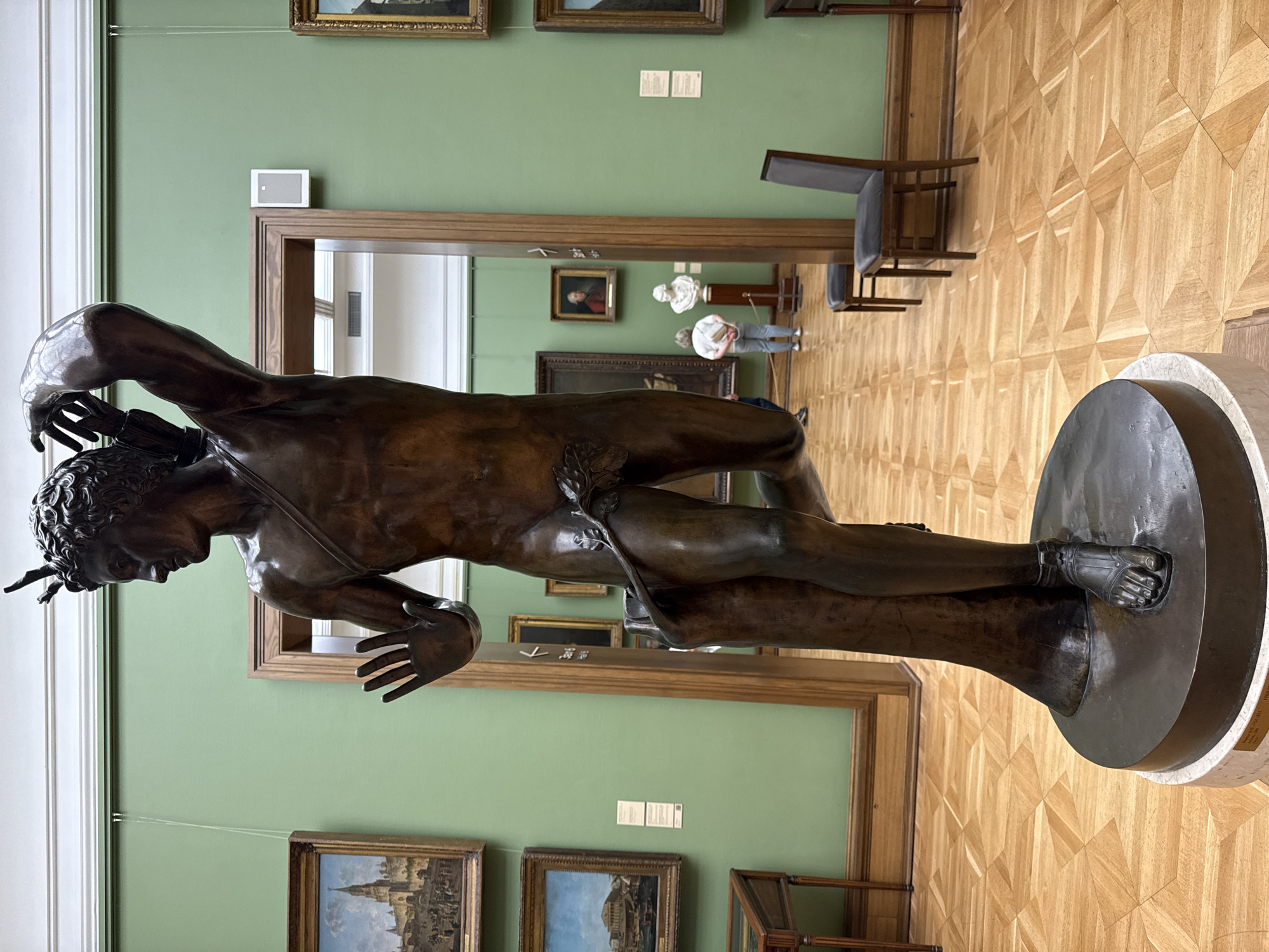 The image shows a bronze statue of a male figure in a museum setting. The statue is on a circular base. The figure appears to be in motion, with one arm reaching out and the other bent. The statue is brown in color.
The museum has green walls and ornate wooden flooring. Paintings hang on the walls. In the background, through a doorway, one can see other museum visitors and displays.
The statue is identified as "Mikhail Kozlovsky, Samson Rending the Lion, 1802". The museum is located in Moscow, Russia.