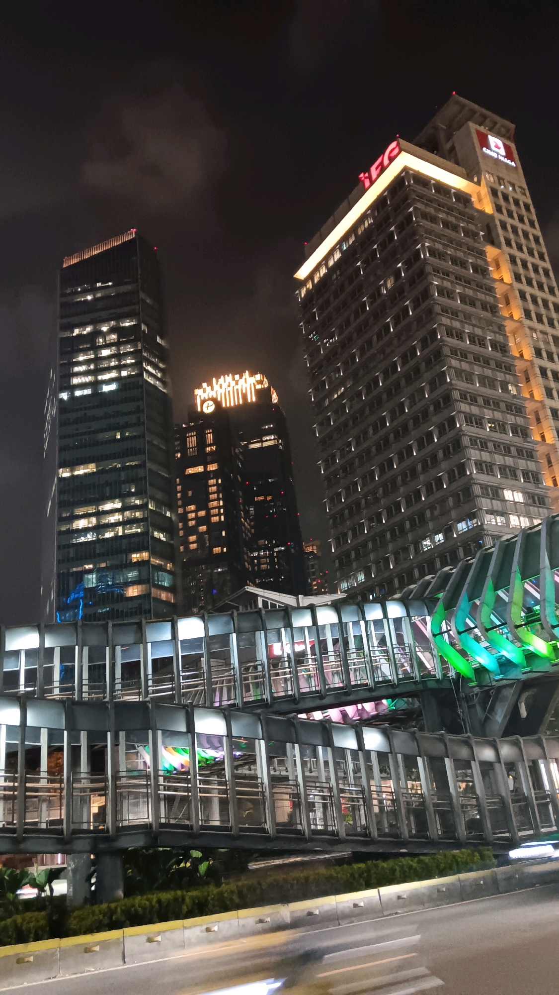 This is a night photograph of the JPO Sudirman pedestrian bridge in Central Jakarta, Indonesia, viewed from a low angle, likely from street level. The bridge, a prominent architectural feature, is illuminated with colorful lights, predominantly green and pink, creating a vibrant contrast against the dark sky and surrounding buildings.
Three tall, modern skyscrapers are visible in the background. The one on the far right has a distinctive design with illuminated horizontal bands and a prominent logo at the top that reads "IFG" in red. Next to it, to the left, is another skyscraper with a lighter facade, also featuring numerous illuminated windows. In the middle ground, a darker skyscraper stands out with an illuminated logo at its peak, appearing as a stylized depiction of a building or structure, with a clock face visible below it.
The JPO Sudirman bridge itself is a multi-level pedestrian walkway with a steel structure and glass enclosures. The metal framework of the bridge is visible, supporting transparent panels that allow glimpses of the colorful lighting within. The lower level of the bridge appears to be more densely constructed, with visible railings and supports.
In the foreground, a street or road is visible, with blurred light trails indicating moving vehicles, suggesting motion and a busy urban environment. A median barrier separates the road from a strip of green foliage, possibly a roadside planter or trees. The overall scene captures the dynamic energy of a modern city at night, with the illuminated bridge serving as a focal point connecting the urban landscape. There are no people clearly visible in the photograph.