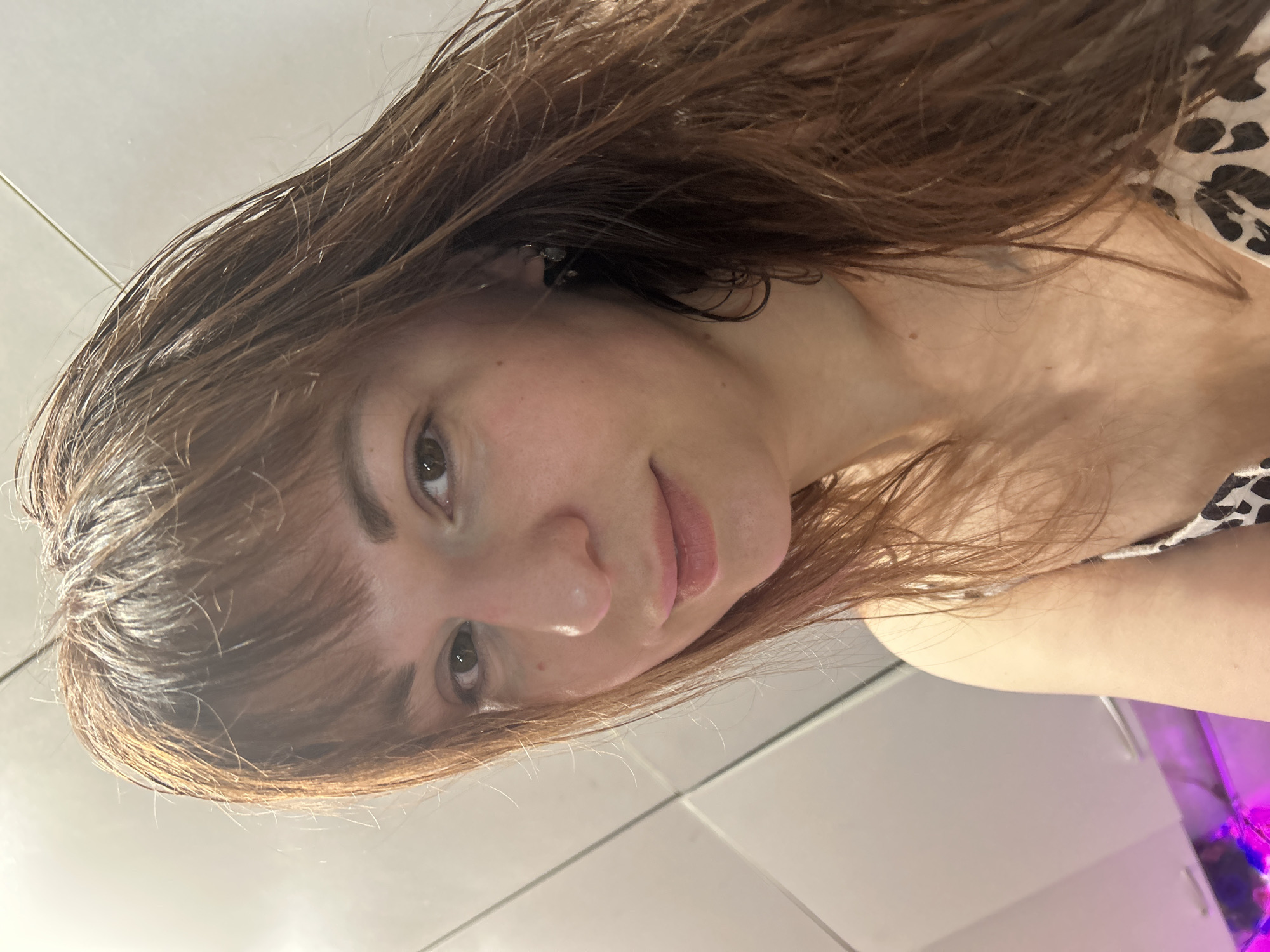 A medium close-up photograph depicts a female subject with light brown, shoulder-length hair and bangs, appearing damp, in an indoor setting identified as Фоки, Russia. She possesses fair skin, light brown eyes, and a slight closed-lip smile while looking directly into the camera. A small, dark earring is visible in her right ear. She is wearing a camisole with a white base and a black or dark brown leopard print pattern. The background consists of light-colored, possibly white, wall panels or tiles exhibiting vertical seams, with overhead kitchen cabinetry visible in the upper left. A diffused purple light source is discernible in the lower left portion of the background, reflecting off an unseen surface.