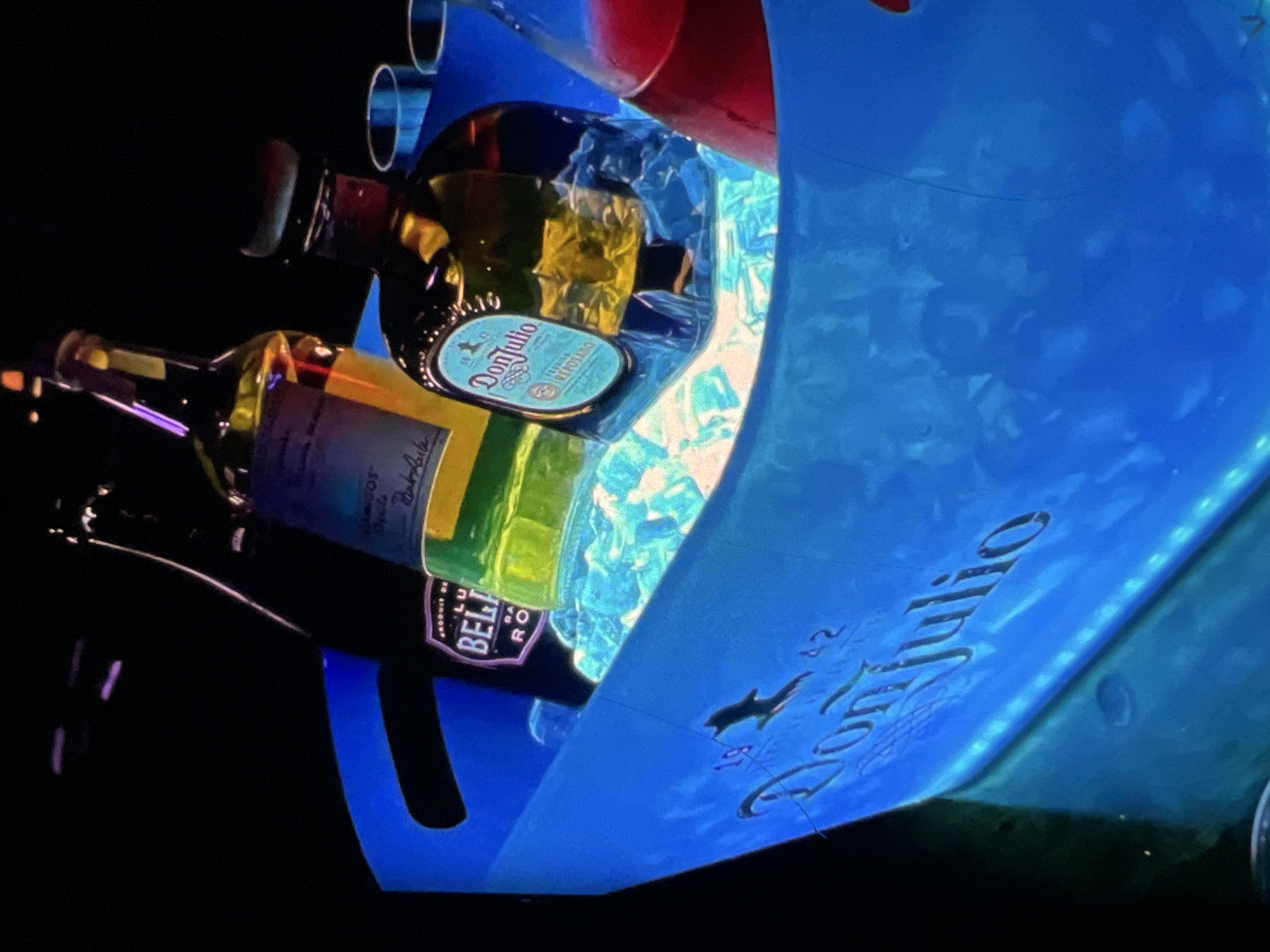 The image shows a blue, illuminated ice bucket filled with ice and several bottles of alcohol. Prominently featured are two bottles of Don Julio tequila, one of which is clearly visible with its distinctive light blue label. There is also a bottle of what appears to be Belvedere vodka with a purple and silver label. A bottle containing a red liquid is partially visible on the right. Two clear shot glasses are placed on the edge of the bucket. The ice bucket itself has the "Don Julio" logo embossed on its side. The background is dark, suggesting an indoor setting, possibly a nightclub or bar in Lagos, Nigeria, given the context provided. The lighting is low, with the blue light from the bucket creating a vibrant glow. There are no people visible in the image.