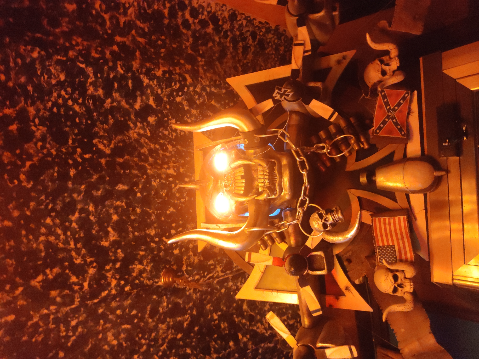 The image shows a close-up, low-angle view of a macabre, horn-adorned skull figure with glowing eyes, possibly a statue or decoration. The figure is adorned with chains, ammunition, and several flags, including an American flag and a Confederate flag. Two smaller skulls with horns are positioned on either side of the flags. The background is a dark, textured surface with an orange, speckled pattern, possibly a wall or ceiling. The lighting is dramatic, with the figure's eyes brightly illuminated. The overall aesthetic is dark and possibly intended to evoke a sense of rebellion or edginess. The location is unknown.