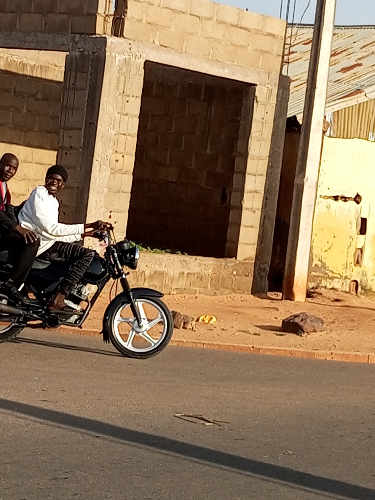 The image depicts two individuals on a motorcycle, riding on a paved road in the city of Gombe, Nigeria. The person in front is driving the motorcycle, wearing a white long-sleeved shirt and dark pants, and a black cap. The person behind them is wearing a red and white shirt and black pants. Both individuals appear to be of African descent. The motorcycle is black and has a silver-colored wheel visible in the foreground. The background shows a building under construction, made of light-colored bricks, with an open doorway. To the right of the building, there is a corrugated metal roof and a utility pole. The ground beside the road is sandy with some scattered debris, including a rock. The lighting suggests it is daytime, possibly late afternoon, given the long shadow cast on the road.