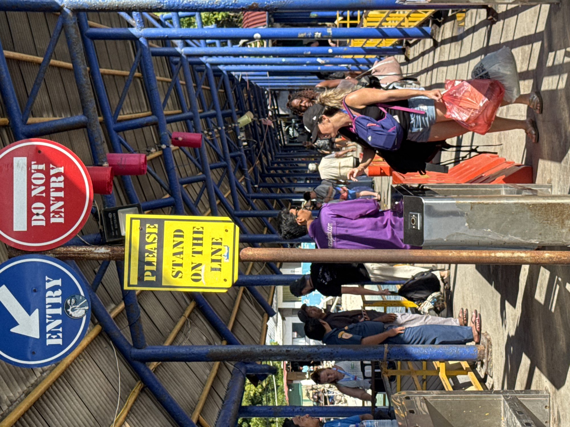 The image displays a bustling outdoor queuing area, likely a pedestrian walkway or entrance to a transportation hub. A prominent overhead structure made of blue metal beams and a light-colored, possibly thatched, roof creates a corridor.
**Subjects:** Numerous people of various ages and attire are forming a queue, primarily facing right. In the foreground of the queue, a person wearing a purple long-sleeved shirt is visible. Further back, a woman in a black top, denim shorts, a black baseball cap, and a purple backpack stands out, holding a translucent red plastic bag. Other individuals are seen throughout the line, some standing, some seated on benches or railings. A person in a dark uniform, possibly security or staff, is also present. The scene includes turnstile-like barriers at the end of the queue.
**Activity or event taking place:** People are waiting in an organized line, likely to pass through an entry point or control gate, suggesting they are boarding a ferry or entering a facility.
**Notable details:** The scene is illuminated by bright sunlight, casting strong, defined shadows on the ground and structure, indicating midday or early afternoon. The weather is clear and sunny. Small red cylindrical objects are suspended from the blue overhead structure. The overall atmosphere suggests a busy public access point in a warm climate.
**Any visible text:**
- A circular red sign with a white horizontal bar and white text reads "DO NOT ENTRY".
- A circular blue sign with a white arrow pointing right and white text reads "ENTRY".
- A rectangular yellow sign with black text reads "PLEASE STAND ON THE LINE".
- The text "easylook.com" is visible on the purple shirt of one person in the queue.