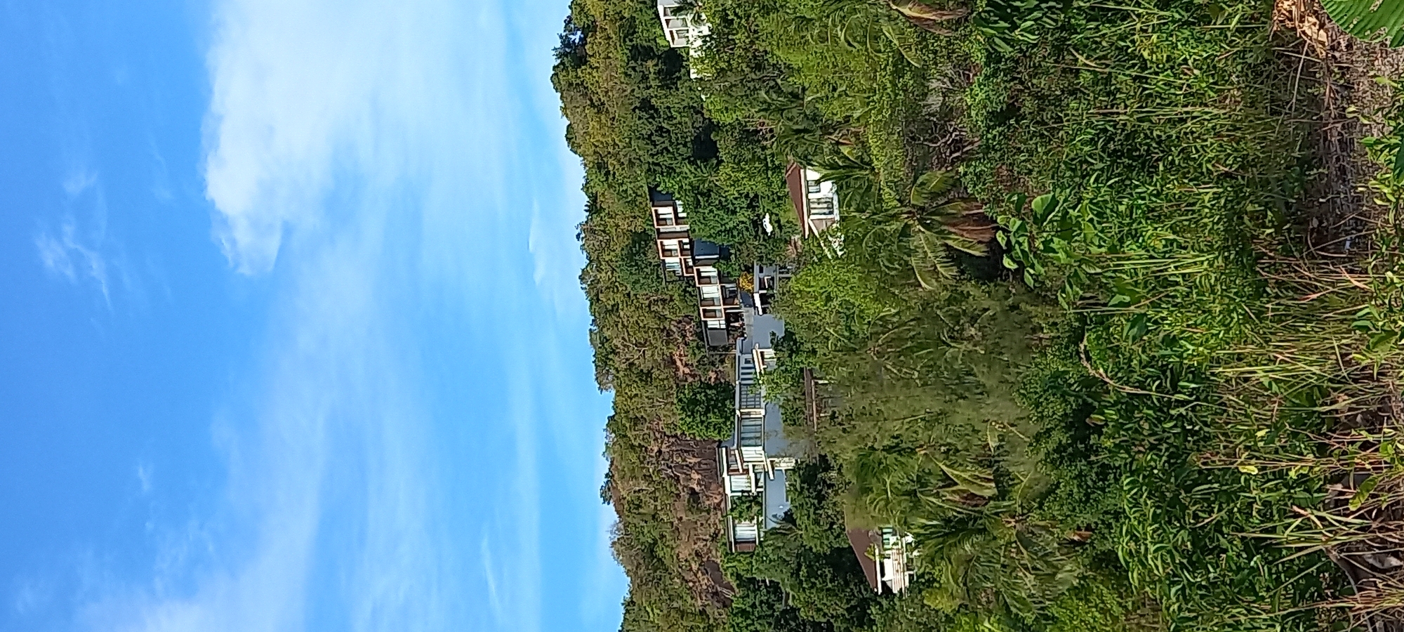 The photograph captures a coastal scene in Baan Wang Ta Kien, Thailand. A series of structures, possibly residential units or resort accommodations, are nestled into the dense tropical vegetation along the shoreline. The buildings feature a modern architectural style, with clean lines and what appear to be large windows or balconies. The vegetation is lush and varied, including tall trees and dense undergrowth, typical of a tropical environment. The sky is blue with some wisps of clouds.