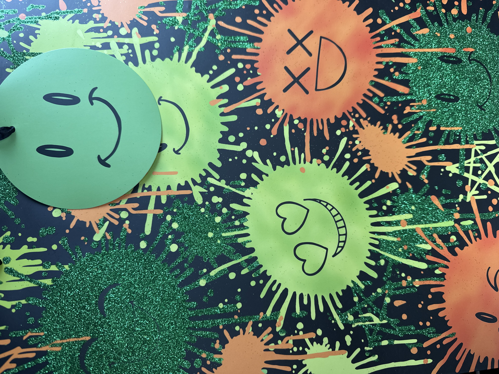 A close-up photograph shows a vibrant, illustrative scene on a dark background, possibly black. A bright green circular object with cartoon-like eyes and a curved mouth sits in the upper left corner. To the right of this object, there are splattered designs in bright shades of green and orange. These splatters are arranged in circular patterns, some adorned with cartoonish faces or expressions: one featuring an "XD" expression, and another incorporating heart-shaped eyes. The background has what appears to be glitter or textured paint in a deep green shade, adding a sparkling effect. The environment is artificial and seems to be a surface or backdrop rather than a real location. There is no indication of Kuta Selatan, Indonesia, in the visual elements of the image.