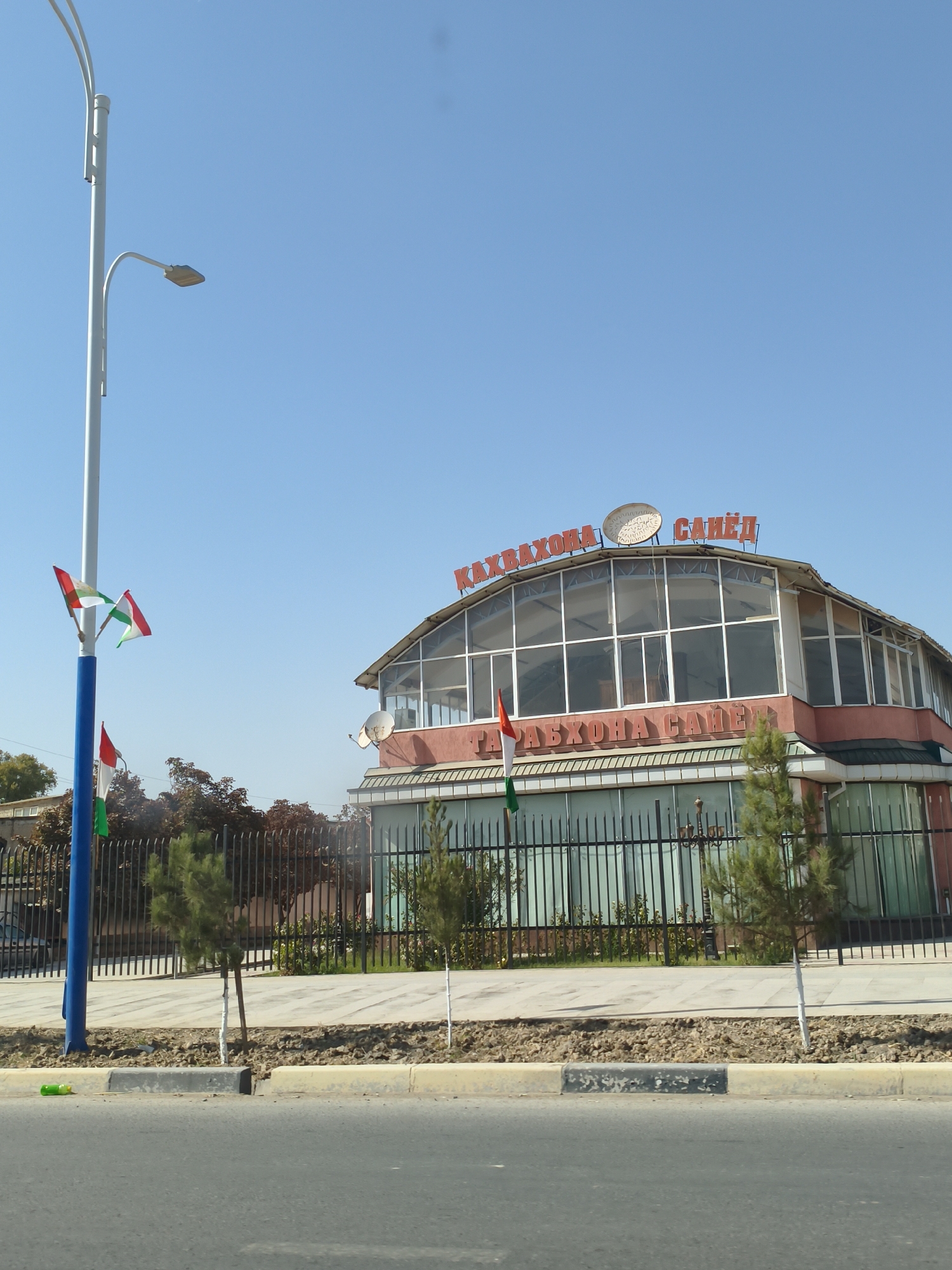 The image captures a daytime street view in Bustonqala, Tajikistan, featuring a building with a distinctive curved glass facade. Red lettering on the building's roof reads "КАХВАХОНА САИЁД" (Kahvakhona Saeiod), suggesting it is a cafe or restaurant. Two satellite dishes are mounted on the roof. A tall street light pole stands to the left, adorned with two flags of Tajikistan. The building is bordered by a black metal fence, behind which are some trees and a landscaped area. A paved sidewalk runs in front of the fence, and a road is visible in the foreground. The sky is clear and blue. A green plastic bottle is lying on the roadside curb.
