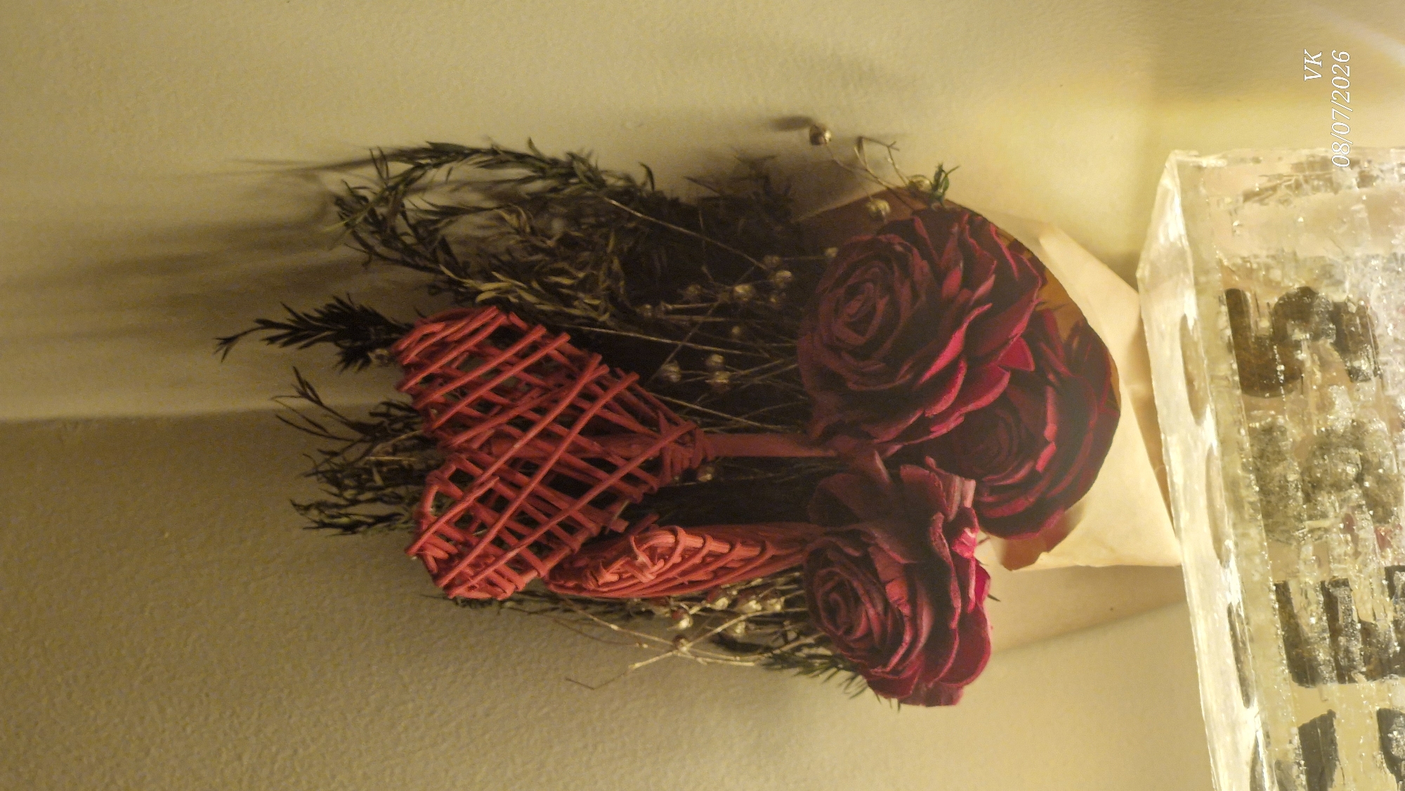 This is a close-up, still life photograph of a bouquet of dark red roses. Two red, woven heart-shaped ornaments are positioned above the roses, with one slightly behind and to the left of the other. The bouquet is arranged in a light-colored paper cone or wrapping, and sits on top of a clear, rectangular object that appears to be made of ice or glass, with some dark objects embedded within it. The background is a textured, light-colored wall, with a shadow cast upon it from the bouquet. The overall lighting is warm and somewhat dim. There is text in the lower right corner that reads "VK 08/07/2026". There are no people or animals visible in the image. The scene does not provide any specific indicators of its location within the city of Vrindavan, India.