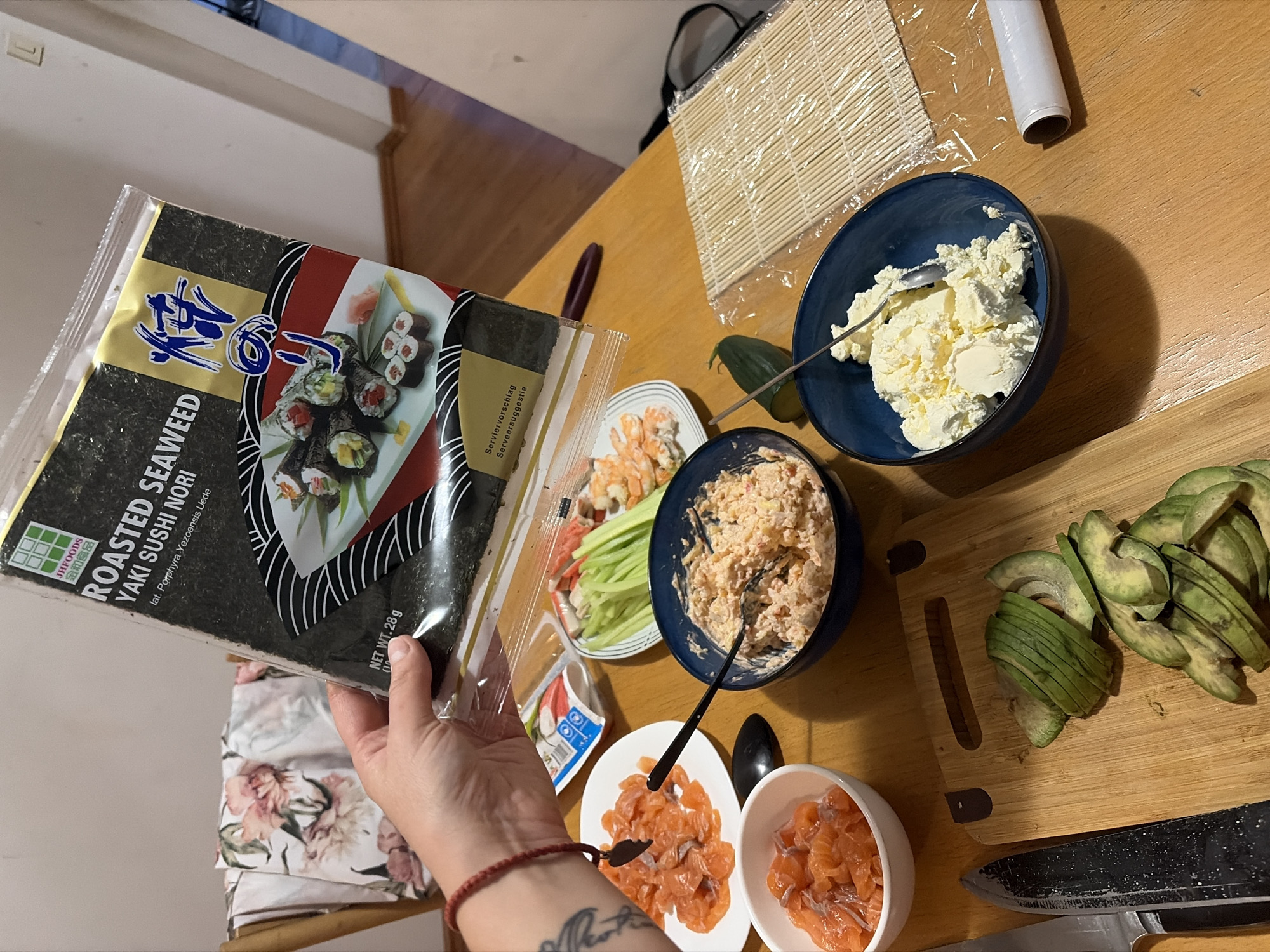 The image depicts a wooden table laden with ingredients for preparing sushi. In the foreground, a bamboo sushi rolling mat is visible, next to a roll of plastic wrap. A blue bowl contains a white, creamy substance, possibly cream cheese, and another bowl holds a mixture of pinkish ingredients, possibly crab or salmon mixed with mayonnaise. Sliced salmon is arranged on a white plate and in a smaller white bowl. Sliced avocado is placed on a wooden cutting board. There are also some vegetables, including cucumber, and what appear to be imitation crab sticks and cooked shrimp on a plate. A package of "Roasted Seaweed Yaki Sushi Nori" is being held by a hand in the upper left corner of the image. The scene suggests the preparation of sushi rolls. The background shows a wall and wooden flooring, hinting at an indoor setting. There is no specific visual information in the image that allows for the determination of the location being Budva, Montenegro.