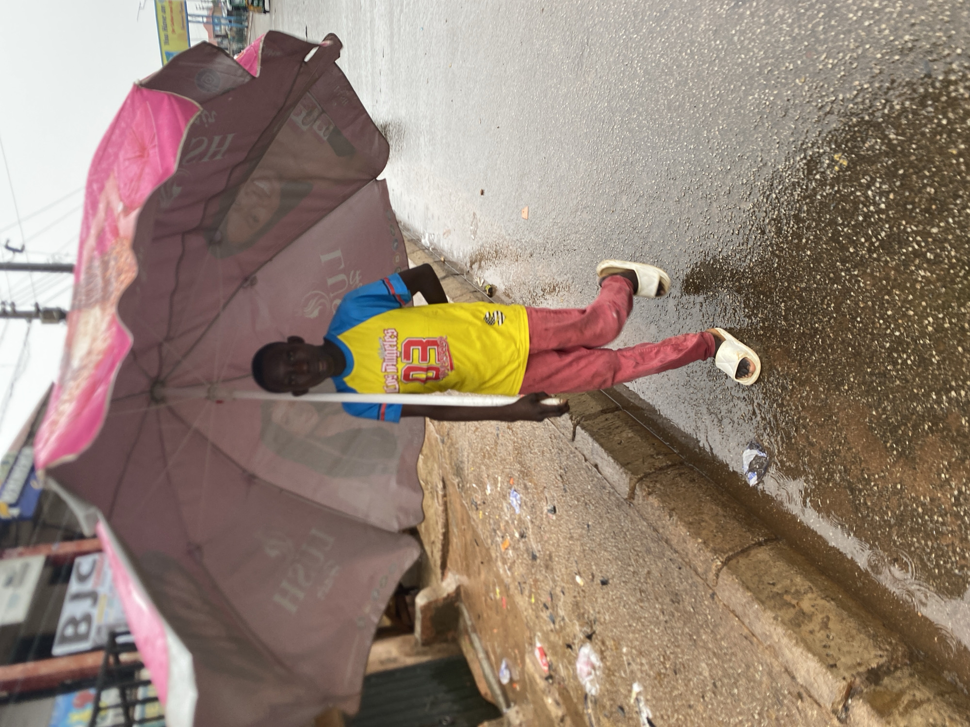 A young person stands under a large, pink umbrella on a wet street. The person wears a yellow shirt with blue sleeves and red pants, along with white sandals. The street is paved and wet, with visible puddles and rain droplets. The curb is lined with litter. The umbrella is branded with commercial images. Background signage hints at a commercial area in Bukuru, Nigeria.