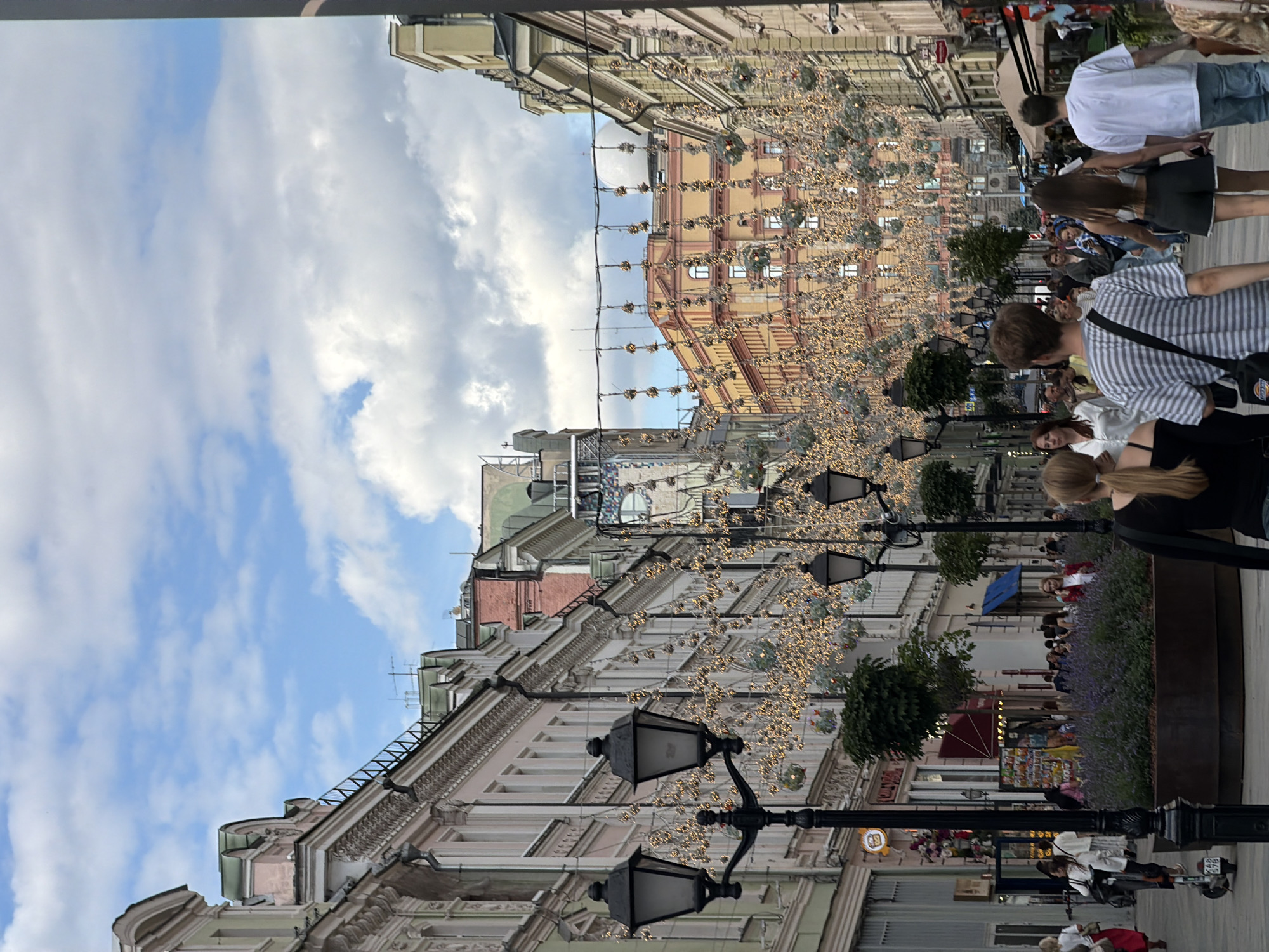 The image captures a street scene in Moscow, Russia, likely during daytime given the bright sky and visible clouds. The perspective is tilted, giving a slightly disorienting but dynamic view of the urban environment.
The foreground features several ornate black lampposts, some with visible light fixtures. Beyond these, the scene opens up to a bustling street lined with multi-story buildings. These buildings display varied architectural styles, with some featuring classical detailing like decorative facades and arched windows, while others appear more modern. Festive lights, strung across the street between the buildings, suggest a special occasion or seasonal decoration, perhaps for a holiday or festival.
Crowds of people are visible, walking along the sidewalks and in the street. They appear to be casually strolling, shopping, or simply enjoying the atmosphere. Their attire suggests a temperate climate. The presence of trees and planters with purple flowers adds greenery and color to the urban landscape, breaking up the more monolithic architectural elements.
Further down the street, more buildings are visible, some with awnings and shop fronts. The general ambiance seems lively and active, indicative of a popular public space. The sky is a mix of blue sky and white clouds, suggesting fair weather. The image conveys a sense of urban vibrancy and community gathering. No specific text is clearly legible in the image.