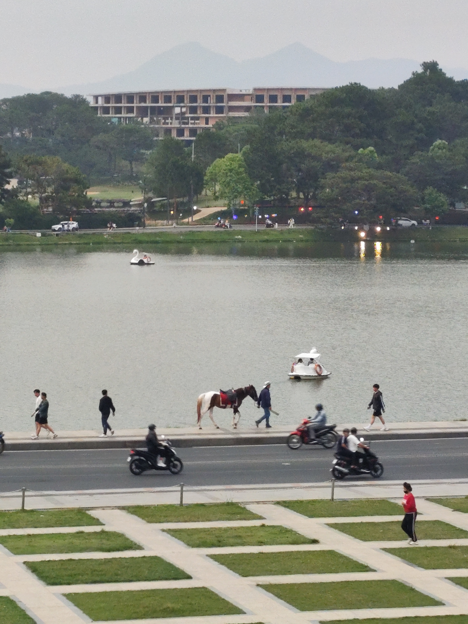 This image captures a multi-layered outdoor scene around a lake in Da Lat, Vietnam, under an overcast sky.
In the foreground, a paved area with an alternating pattern of grass squares and light-colored paths is visible. A person in a red top and dark pants walks towards the right across this area.
Above this, a road runs parallel to the lake, bustling with activity. Several individuals are seen riding motorcycles; at least four motorcycles are distinguishable, some carrying passengers. Adjacent to the road, a pathway along the lake is used by pedestrians. A group of three people walks left, while another person walks right. Notably, a brown and white horse, saddled, is being led by a person along this pathway.
The calm waters of the lake reflect the soft light from the sky and the glowing headlights of vehicles on the opposite bank. Two paddle boats are present on the water: one distinct swan-shaped vessel and another smaller white boat carrying occupants.
The far bank of the lake is densely covered with green trees. Through the trees, parts of another road are visible with a white car and what appear to be streetlights emitting various colors. Further in the background, a large, multi-story concrete building with numerous open window frames, appearing unfinished or derelict, stands prominently on a tree-covered hill. Distant, hazy mountains are visible beyond the hills, blending into the soft, light grey sky.
The scene suggests a leisurely atmosphere, possibly late afternoon or a very cloudy day given the diffused lighting and the active streetlights. There is no visible text in the image.