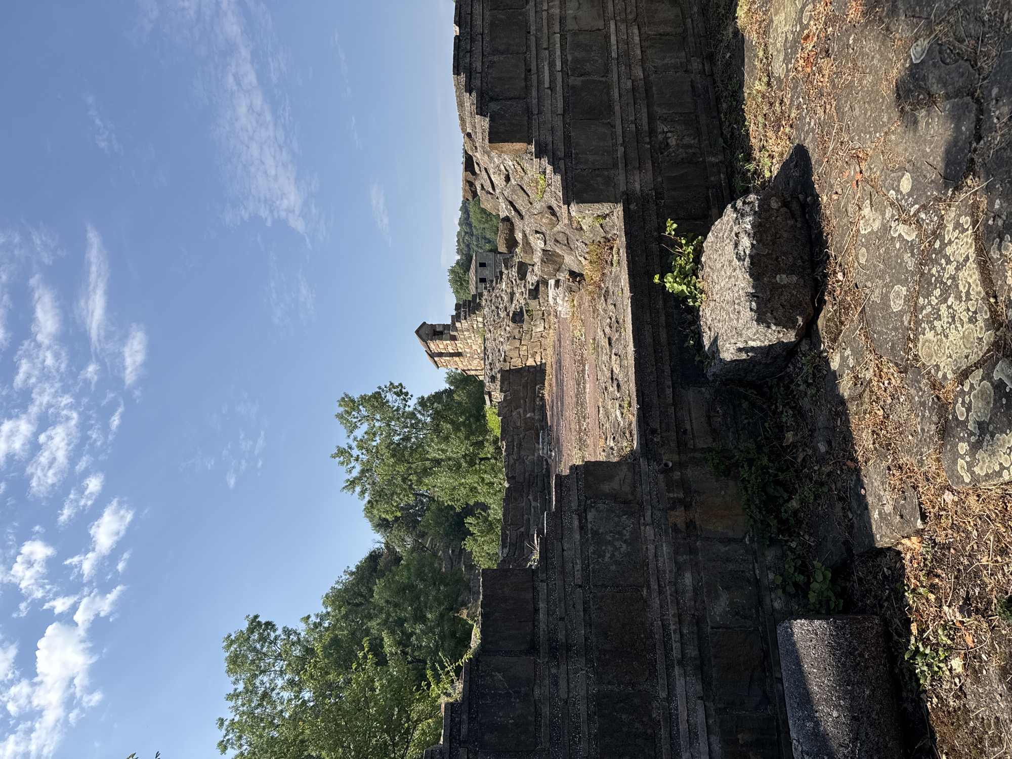 The image shows the ruins of an ancient structure, likely a fortress or a complex of buildings, under a bright blue sky with scattered white clouds. The ruins are constructed from large, dark stones, forming walls, steps, and platforms. In the background, lush green trees cover a hillside. A small, two-story stone building with a pitched roof sits atop the ruins in the mid-ground. Another similar, smaller structure is visible to its right. The ground is a mixture of stone paving and dry earth, with some green vegetation growing amongst the stones. The scene suggests a historical site in an outdoor, natural setting. This location is the Tsarevets Fortress in Veliko Tarnovo, Bulgaria. There are no people visible in the image.