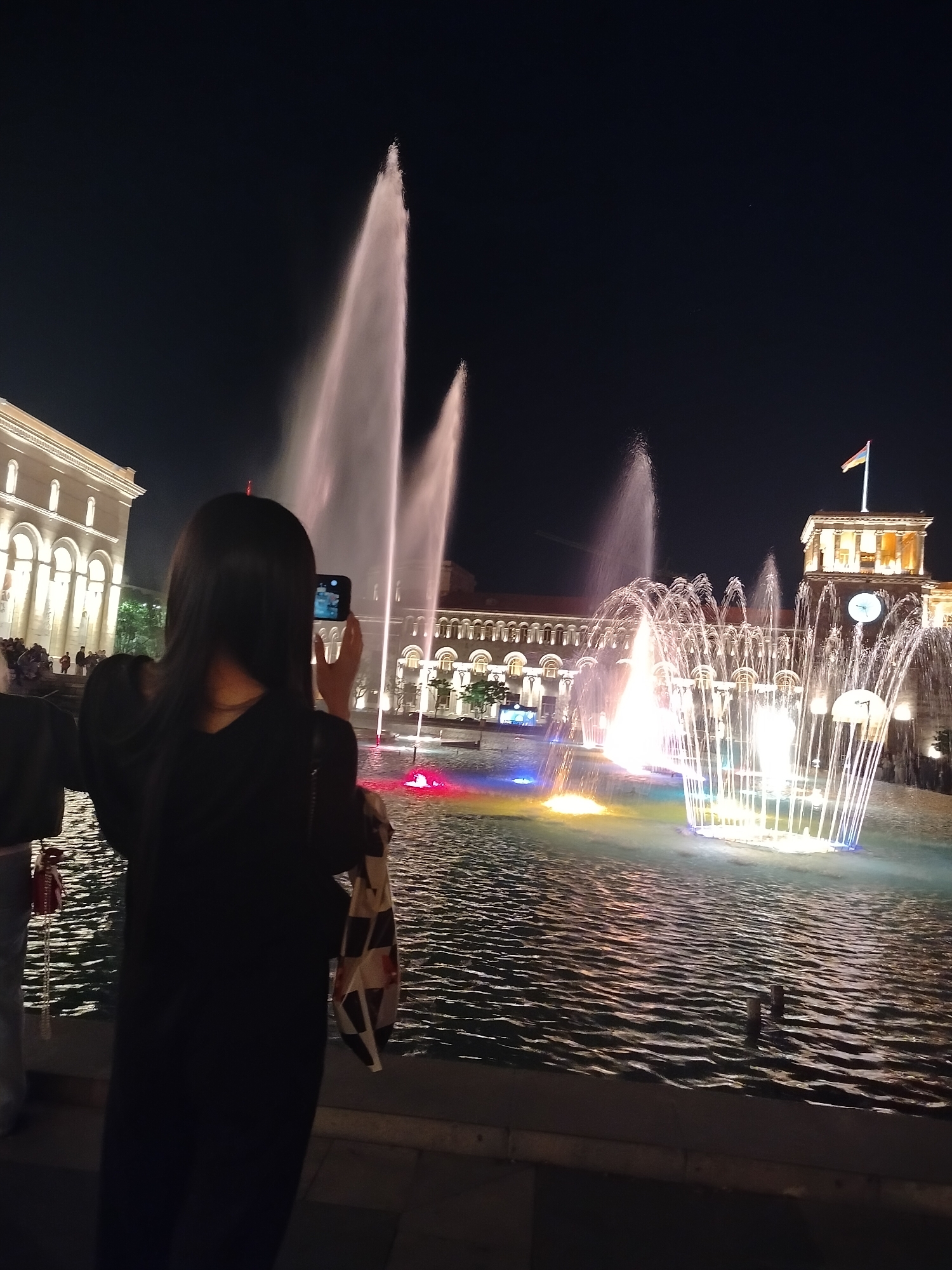 At night, a person with long, dark hair stands with their back facing the viewer, holding a smartphone to capture a photo or video of a large, illuminated fountain. The fountain features multiple water jets of varying heights and formations, with vibrant colors shining through the water. Beyond the fountain, illuminated buildings are visible. A tower with a clock face and a flag on top is also visible. People are seen in the background near the building on the left. The location is Republic Square in Yerevan, Armenia. The fountain is the main focal point, reflecting light onto the rippling water surface.