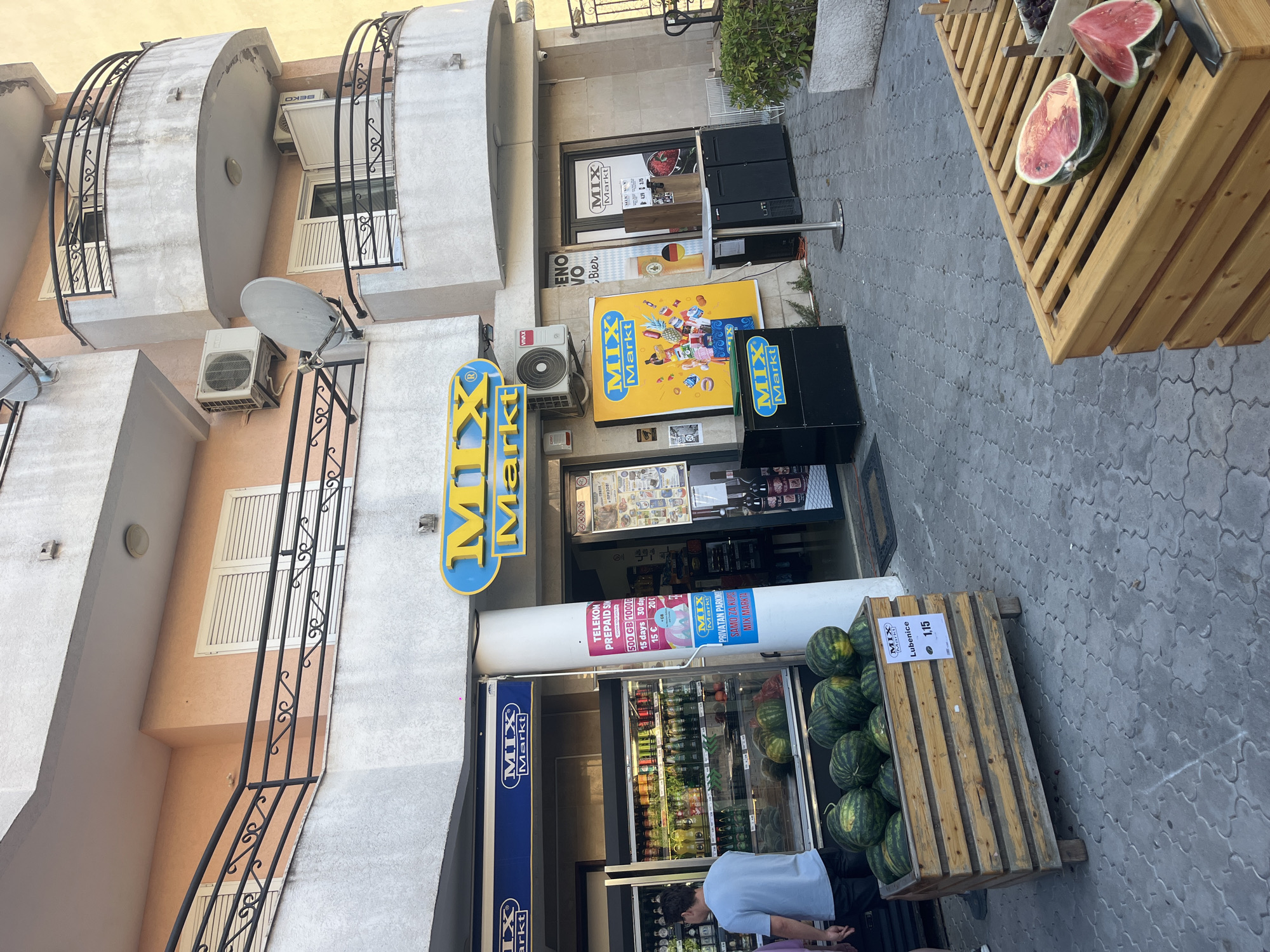 The image shows the exterior of a "MIX Markt" store. In front of the store, there is a display of watermelons in a wooden crate. One of the watermelons has a price tag of "1.15". To the right of the watermelon display, there is another wooden display with cut watermelons. A person is visible on the left side of the image, standing in front of a refrigerator filled with drinks. The store has a sign with "MIX Markt" in yellow and blue. There are also several posters and advertisements displayed on the store's facade. The environment appears to be an urban setting with buildings and pavement.