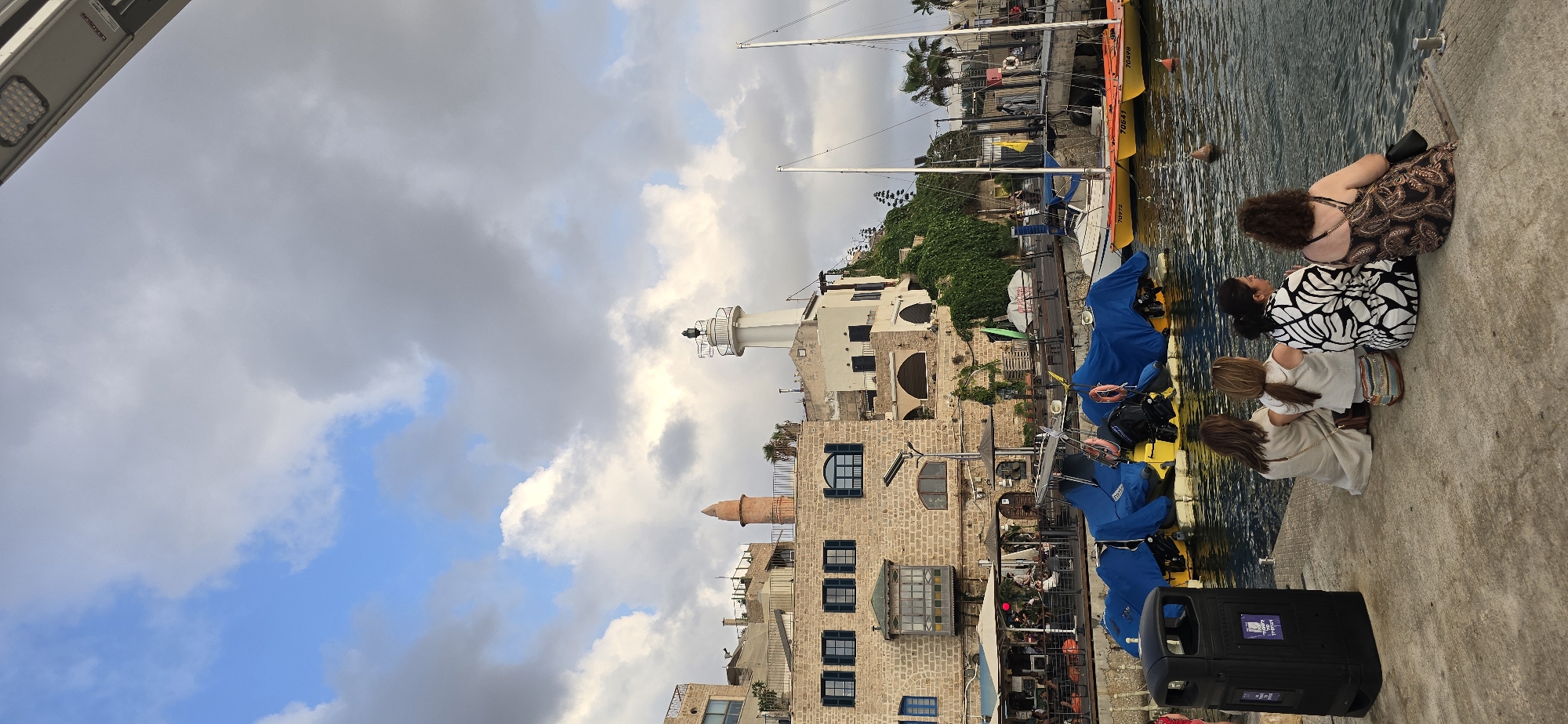 The image depicts a waterfront scene in Jaffa, an ancient port city that is part of Tel Aviv-Yafo, Israel. In the foreground, four women are seated on a concrete pier, facing the water. They are dressed casually and appear to be observing the harbor.
To their left, a black trash receptacle stands prominently. The water in the harbor is calm, reflecting the sky and the surrounding structures. Several boats are docked, some covered with blue tarps, while others, particularly yellow dinghies with identifying numbers (e.g., 70972, 70641, 70498), are visible.
In the mid-ground, a stone building with dark-shuttered windows and a decorative balcony with stained glass is visible. Adjacent to this building are more stone structures with arched windows, some adorned with greenery cascading down the walls. A distinctive, conical minaret rises from one of the buildings, and a white lighthouse with a lantern room stands on a hill overlooking the port. The sky above is partly cloudy with patches of blue sky visible. The overall impression is a tranquil day at a historic harbor.