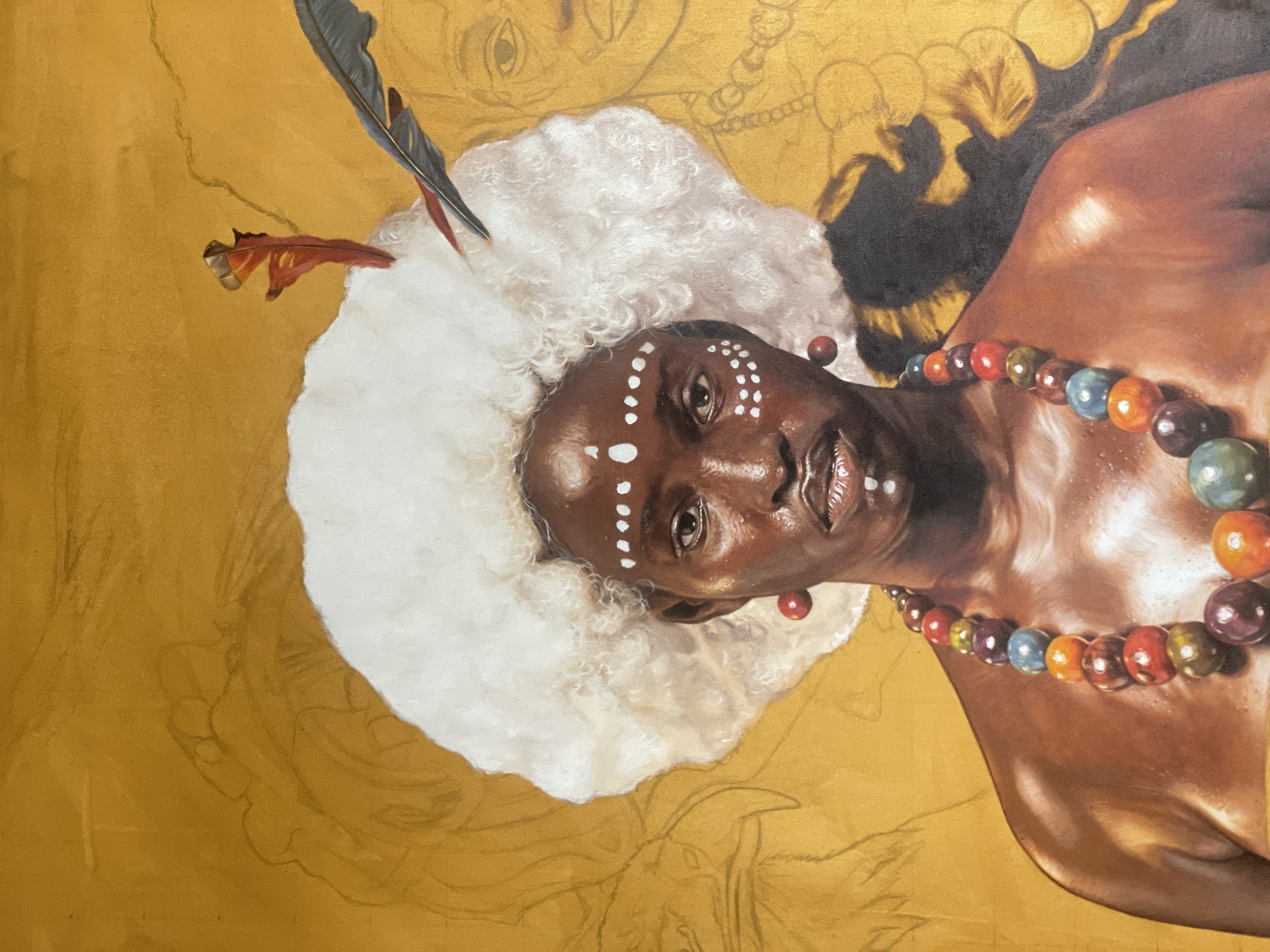 A portrait of a dark-skinned person with white dots on their forehead and cheekbones. They wear a large white, ruffled headpiece adorned with a feather and a red ornament, and a necklace of colorful spherical beads. The background is a gold color. The location in the city of Ado, Nigeria is not determinable from the image.
