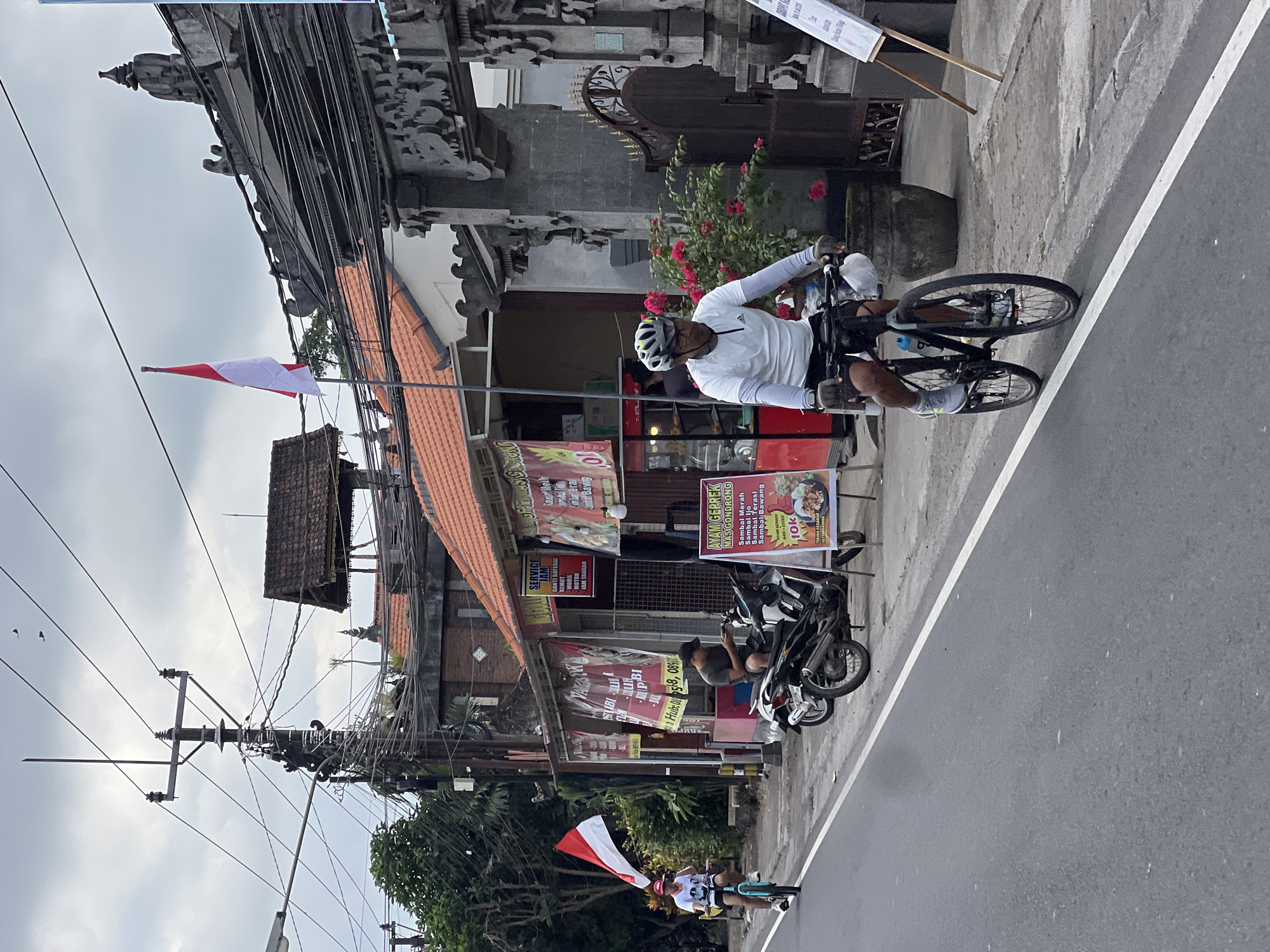 In Abiansemal, Indonesia, on a street with a white dividing line, two individuals are cycling. A man in a white long-sleeved shirt and helmet rides a black bicycle in the foreground, while another cyclist, wearing a red cap, rides a blue bicycle further down the road. The street is bordered by shops and buildings with red tile roofs, ornate stone gates, and numerous overhead electrical wires. Several motorcycles are parked along the roadside, and various banners and signs are displayed outside the businesses. A red and white Indonesian flag is visible flying from a pole. The sky is overcast, and the environment appears to be a commercial area within the town.