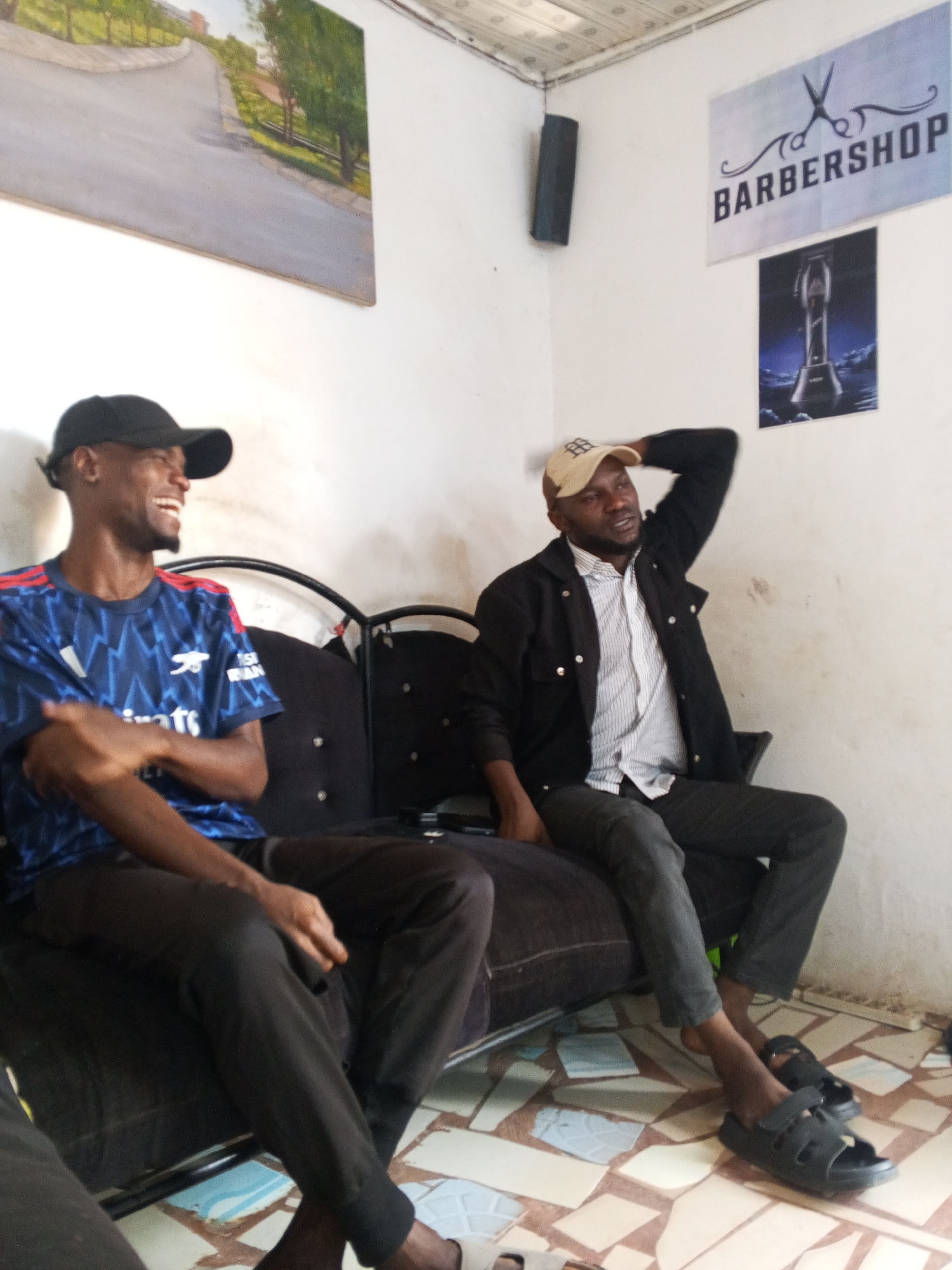 Two men are seated on a dark-colored couch in what appears to be a barbershop in Gwong, Nigeria. The man on the left wears a black baseball cap and an Arsenal soccer jersey. He is smiling. The man on the right wears a beige baseball cap, a white striped shirt, and a black jacket. He has his right arm raised, touching his head. The barbershop's wall features a "BARBERSHOP" sign, an advertisement for a hair clipper, and a painting. The floor has a mosaic tile pattern.