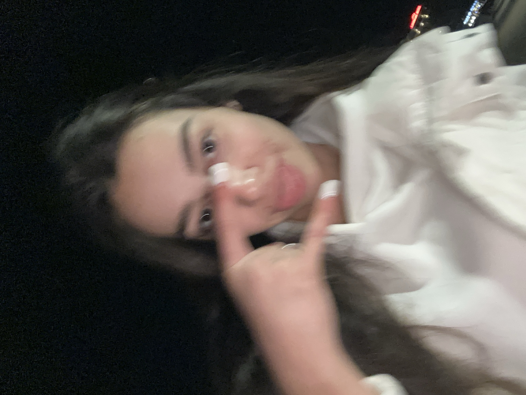 A blurry, close-up photograph shows a young woman with long, dark hair. She is making a hand gesture with her index and pinky fingers extended and her middle and ring fingers folded down. She wears white nail polish on her fingers. She is wearing a white garment. The background is dark. A few blurred lights are visible in the upper right. The photograph does not provide enough information to determine a specific location in Plovdiv, Bulgaria.