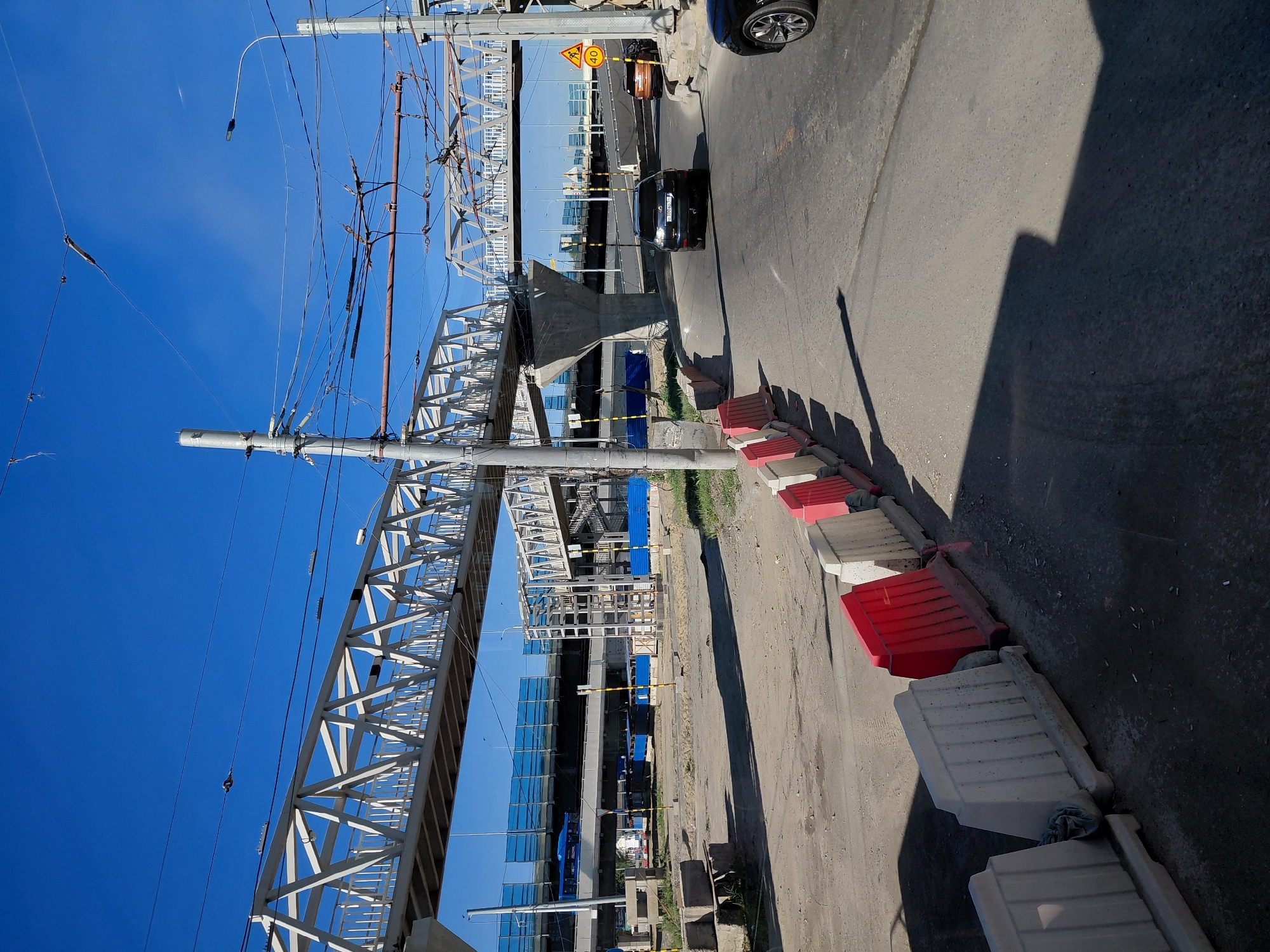 In an outdoor urban environment under a clear blue sky, a pedestrian overpass constructed of white steel lattice spans across a roadway. The overpass is supported by concrete pillars. The surrounding area appears to be a construction zone, with concrete barriers, some red and some white, lined up along the side of the road. Electrical wires and poles are prominent in the scene, with a street lamp visible to the right. A black SUV is driving on the road, and an orange car is parked further down. In the background, another elevated road with blue barriers is visible, and a blue bus is seen on it. The ground is a mix of dirt and asphalt.