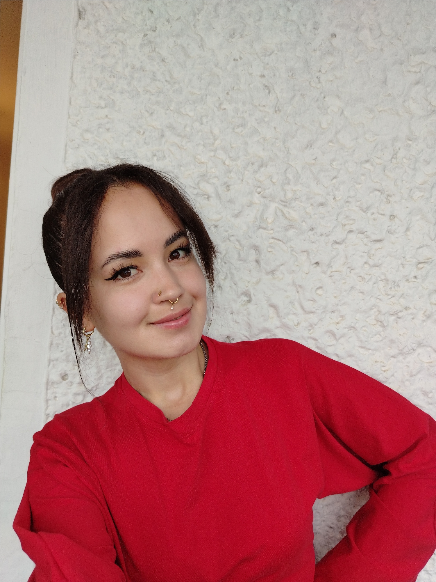 A young woman with dark hair pulled back in a bun, bangs framing her face, and light brown eyes is pictured smiling. She is wearing a bright red long-sleeved shirt. She has a nose ring with a small diamond and a septum ring with a dangling pearl. Her right ear has multiple piercings, including one with a pearl drop earring. She is positioned in front of a textured, pale gray wall. The image is a medium shot, focusing on her upper body and face. She appears to be indoors or in a covered outdoor space, as indicated by the wall texture and indirect lighting. The image does not contain information about the city of Noviy, Russia.