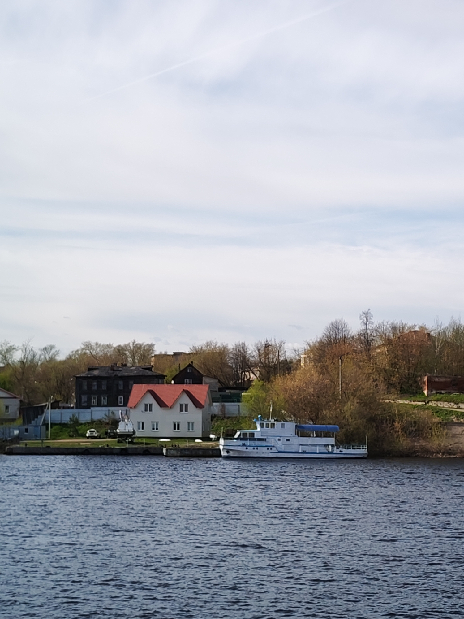 The image captures a tranquil scene in Kimry, Russia. A body of water, presumably the Volga River or one of its tributaries, dominates the foreground with dark, rippled water. Along the shoreline, a small white and blue passenger boat is docked, suggesting the river is used for local transport or tourism.
On the land, a mix of structures is visible. There is a white house with a distinctive red pitched roof and multiple windows that add to the architectural detail. To the left of the house is a dark-colored multi-story building. The trees surrounding the buildings are sparse with few leaves, suggesting early spring or late autumn. A car is parked near the white house.
The sky is overcast with thin clouds, which create a soft, diffused light across the scene. The overall impression is one of a calm, settled environment within a typical Russian townscape. The absence of people in the shot adds to the quiet, contemplative feel of the image.