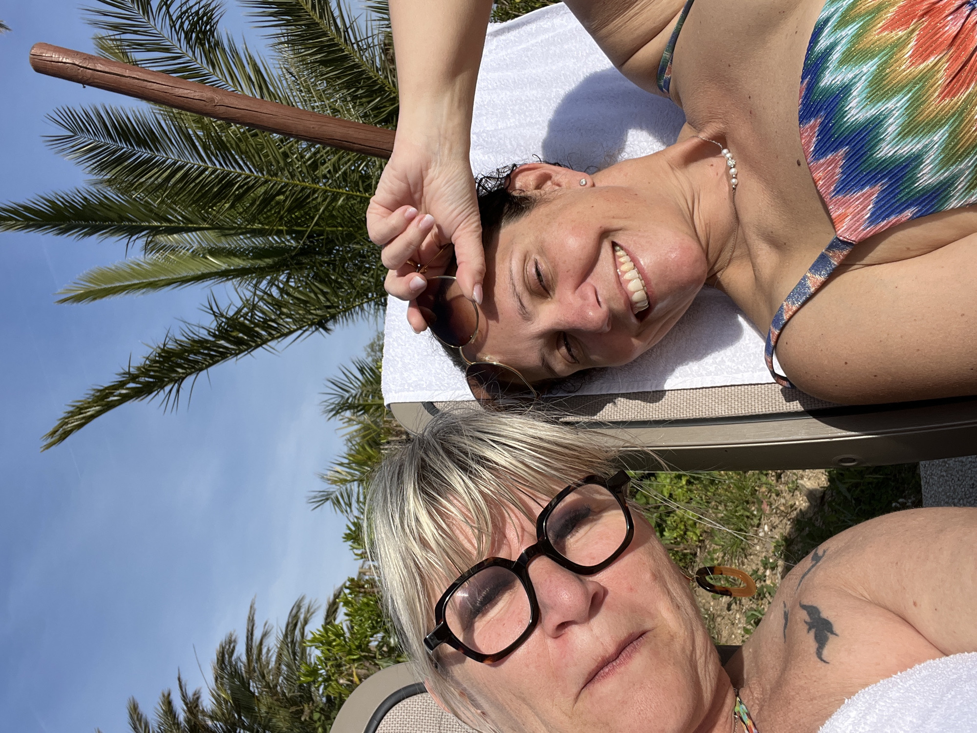 Two women are lying on a lounge chair outdoors. One woman is wearing a multicolored bathing suit and has her hand raised near her sunglasses. She is smiling. The other woman is wearing glasses and has a tattoo on her shoulder. The location appears to be Grimaud, France based on the palm trees and other vegetation. The image is taken from a high angle looking down on the women.
