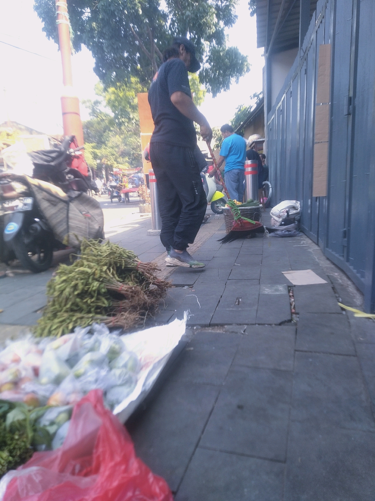 The image depicts an outdoor scene in Cianjur, Indonesia, likely a street or market area. In the foreground, a person wearing a dark t-shirt and dark pants stands with their back to the camera, appearing to be a vendor. In front of them, a large bunch of green leafy vegetables, possibly herbs or greens with roots, lies on the ground, tied with red string. To the left of the vegetables, there are plastic-wrapped produce items, including what appear to be fruits and vegetables, some in a white sheet or bag. Further back, a yellow scooter is parked, and a person in a blue t-shirt and jeans is visible near a basket. A dark metal fence runs along the right side of the frame, separating the sidewalk from an adjacent building or property. The ground is paved with dark square tiles, some of which appear to be broken or uneven. Trees with dense foliage are visible in the background. The lighting suggests it is daytime.