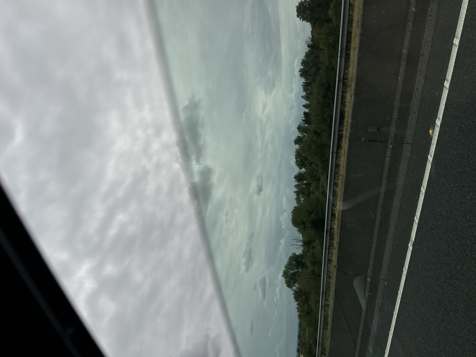 The image captures a view from a vehicle traveling on a highway, likely in or near Ballitore, Ireland, given the context provided. The scene is dominated by a cloudy sky, with a distinct diagonal line cutting across the frame, possibly representing the window of the vehicle or a reflection. Below the sky, a dense line of trees forms a natural barrier. In the foreground, a dark asphalt road with a white lane marking is visible, indicating a roadway. A concrete barrier separates the road from the area with the trees and sky. The overall impression is one of overcast weather and a typical roadside landscape. There are no people or specific actions depicted in the image.