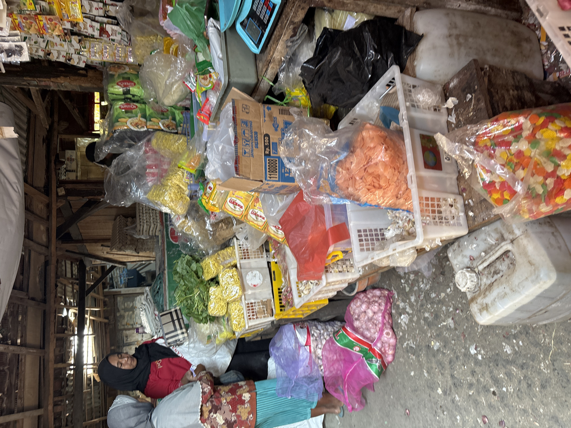 The image depicts an indoor market scene in Tirtomoyo, Indonesia, likely a traditional market given the rustic wooden structure and the variety of goods. In the foreground, two women are visible, one wearing a grey headscarf and a patterned top, the other a black headscarf and a red shirt. The woman in the red shirt appears to be interacting with the woman in the grey headscarf.
The market stall is cluttered with various food items and merchandise. Piles of red onions are prominent, some contained in pink plastic bags. Bunches of green leafy vegetables are also visible. Several plastic containers and baskets hold an assortment of products, including what appear to be dried noodles, packaged snacks, and possibly crackers or chips in orange and multicolored varieties. Bags of rice or other dry goods are stacked in the background, along with numerous packaged food items displayed on shelves. A blue electronic scale is positioned on a wooden surface.
The environment suggests a functional, working market with goods displayed on tables and the floor. The ground is littered with debris, consistent with a busy marketplace. The lighting appears to be a combination of natural light filtering through the open structure and possibly artificial light sources, though none are clearly visible in operation. The overall impression is one of a bustling, localized retail setting.