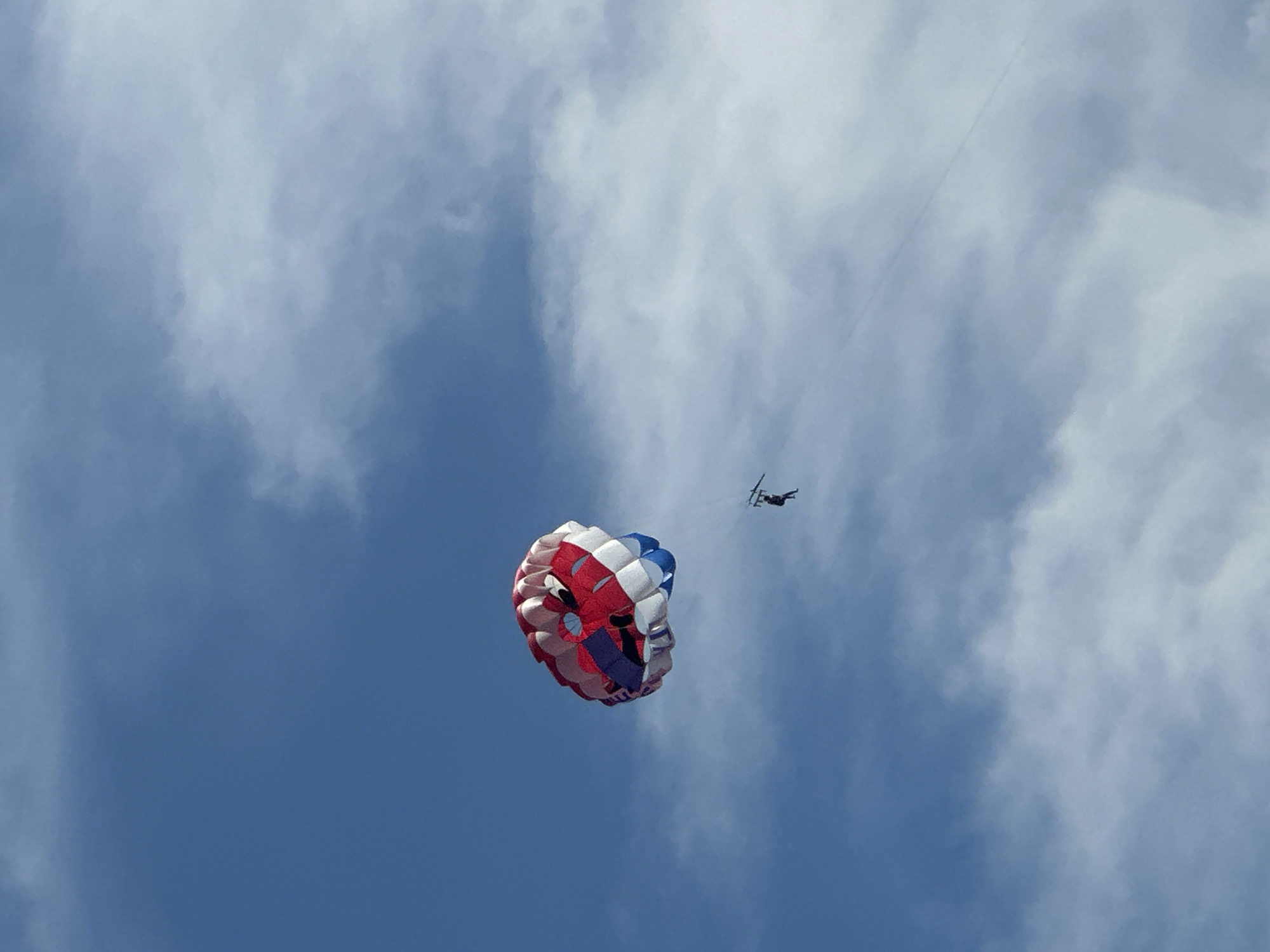 The image captures a parasailer soaring against a bright blue sky with scattered white clouds, suggesting a daytime scene, possibly in Budva, Montenegro. The parasail is a vibrant mix of red, white, and blue. A single person is suspended beneath the parasail, appearing to be enjoying the flight. The perspective is from below, looking upwards at the parasailer and the expanse of the sky. The overall scene evokes a sense of adventure and freedom.