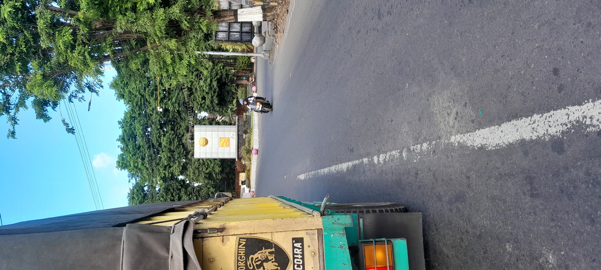 A low-angle, eye-level shot shows a busy street scene in Rogojampi, Indonesia, on a clear, sunny day. The foreground is dominated by the side of a yellow and green truck, with a black tarp covering its load. On the truck's side is a prominent black and gold emblem featuring a bull-like creature, with the word "LAMBORGHINI" above it and "COIRA" below. The truck is on the right side of the road, partially obscuring the view.
The road itself is asphalt, with a white line marking the lane division. In the mid-ground, a person wearing a blue jacket and white helmet is riding a black motorcycle, moving away from the camera. Behind the motorcycle, a modern monument stands in the center of a roundabout. The monument is white, rectangular, and topped with a golden circular emblem, with other golden shapes adorning its sides. Surrounding the monument is greenery, including trees and bushes, and in the distance, more trees line the horizon. The sky is a clear blue, with a few wispy white clouds. The overall impression is one of everyday life in a bustling Indonesian town.