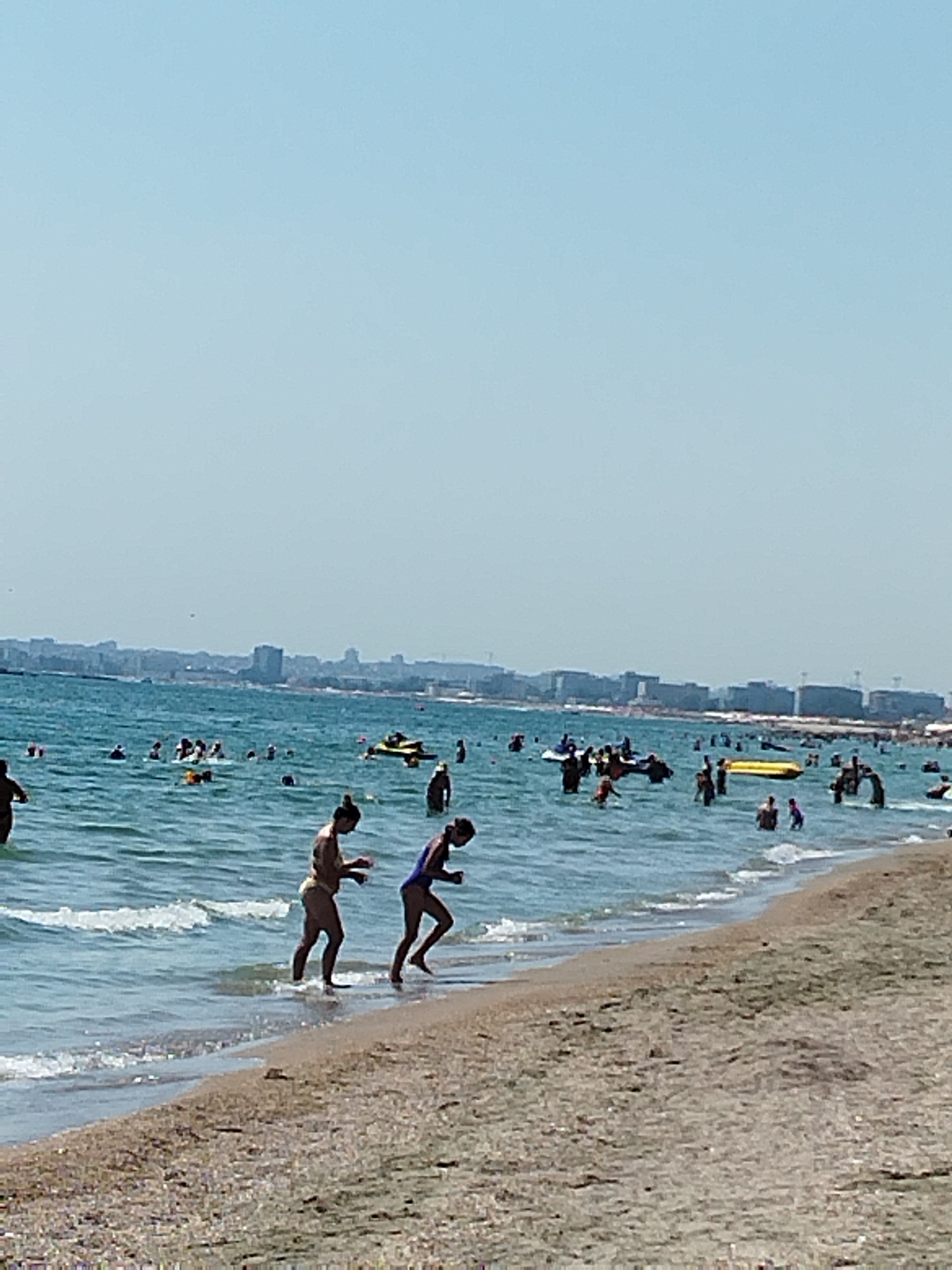 The image depicts a crowded beach scene in Constanța, Romania, under a clear, bright sky. The foreground is dominated by a sandy beach with some scattered shells. In the shallow waters along the shoreline, two women are wading, one in a yellow swimsuit and the other in a blue one. The middle ground is filled with numerous people swimming and standing in the sea. Several inflatable watercraft, including a yellow banana boat and a jet ski, are visible with people on or around them. In the background, a city skyline with buildings and possibly industrial structures stretches along the horizon, indicating the urban setting of Constanța. The sea appears to be calm with small waves lapping at the shore.