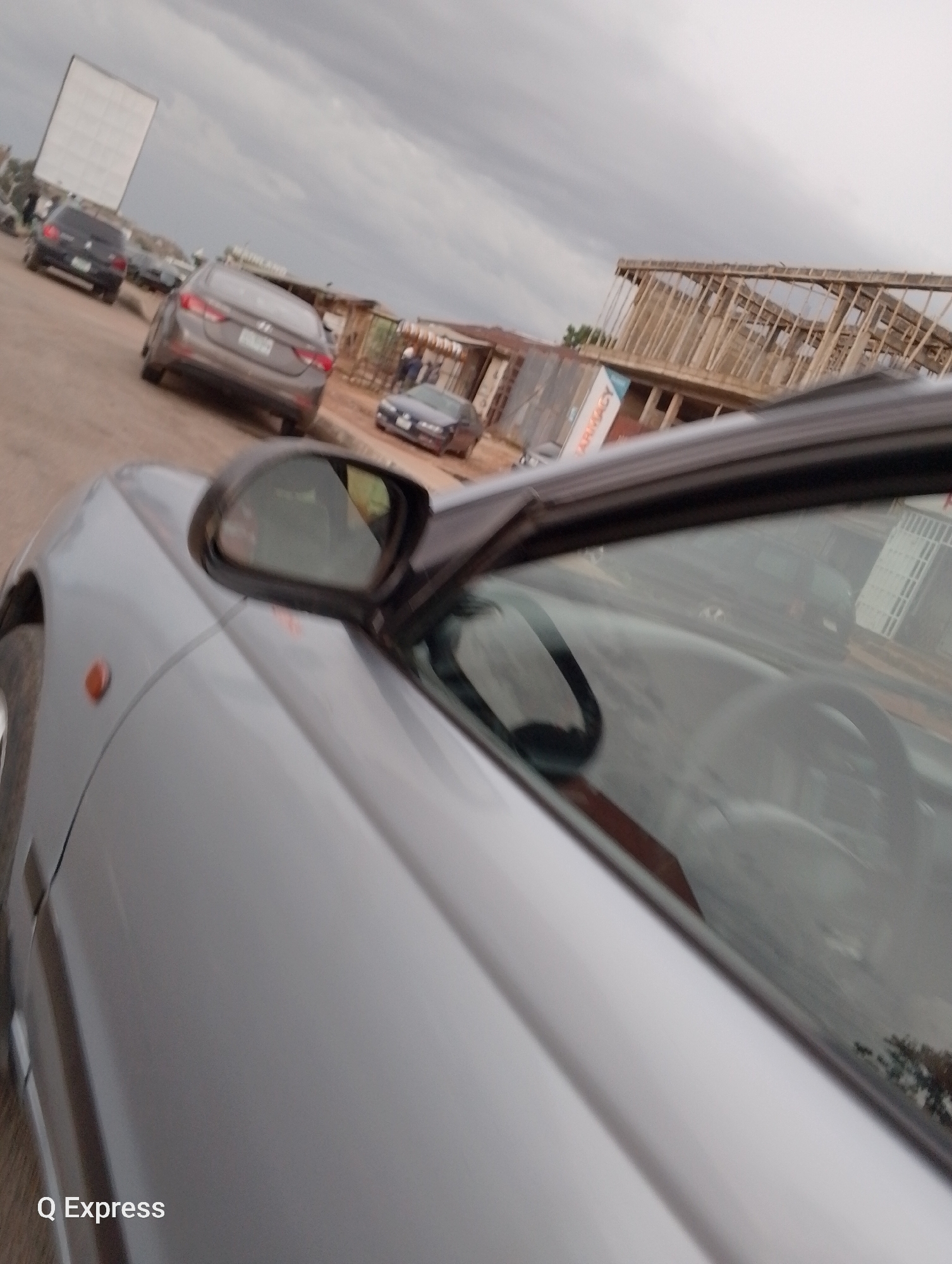 The image shows a street scene in Jos, Nigeria, with several cars and a building under construction. A silver convertible car is in the foreground, with its side mirror visible. Behind it, a gray sedan, a black SUV, and a blue sedan are parked or driving on the road. In the background, a partially constructed building with wooden scaffolding dominates the scene. There is also a large white screen, possibly for advertising or a drive-in cinema, to the left of the frame. The sky is overcast, suggesting cloudy weather. The text "Q Express" is visible in the bottom left corner of the image.