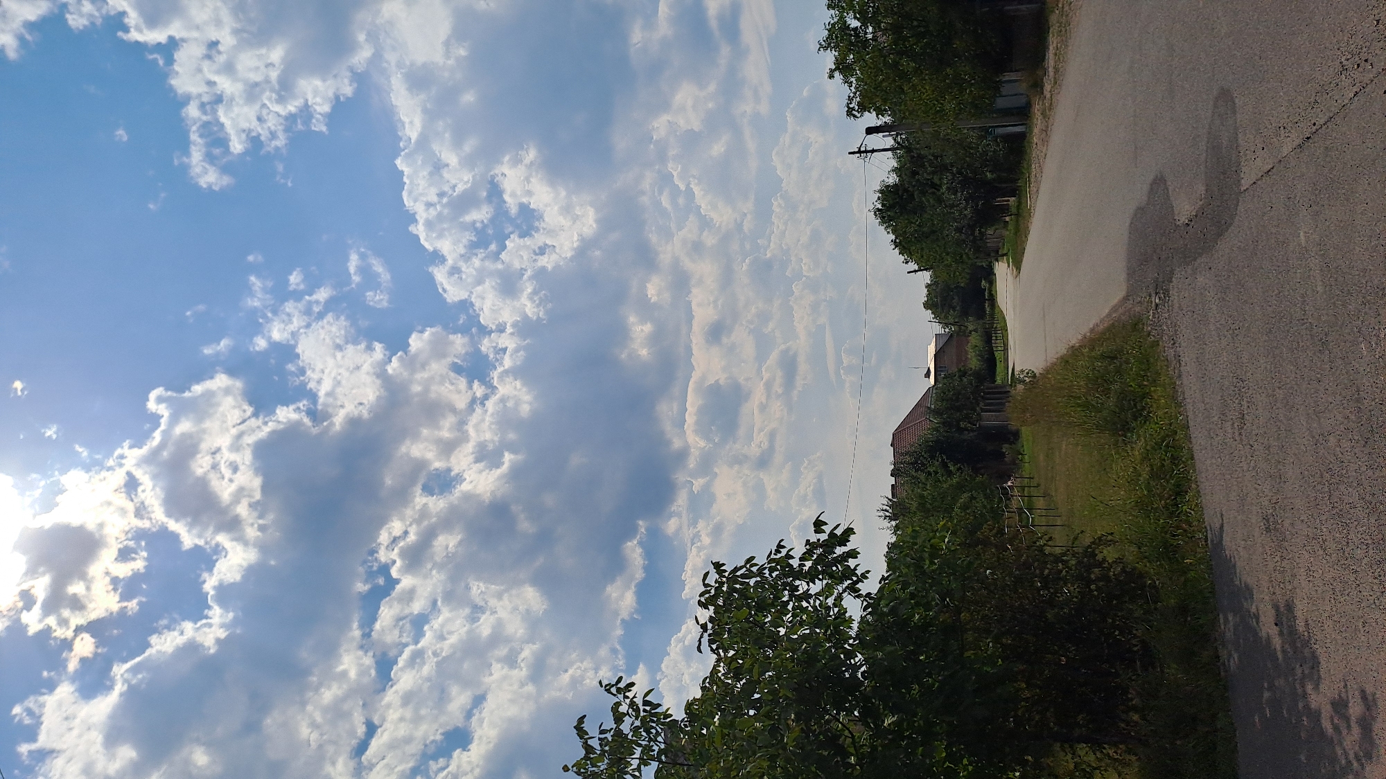 A paved road curves through a semi-rural area with lush green trees and grass on either side. In the distance, residential buildings with red tile roofs are visible. The sky above is a vibrant blue, punctuated by dramatic, white cumulus clouds. The sun is shining brightly, casting dappled shadows on the road. The overall impression is one of a pleasant, sunlit day in a tranquil setting. The image was captured in Kolomyia, Ukraine.