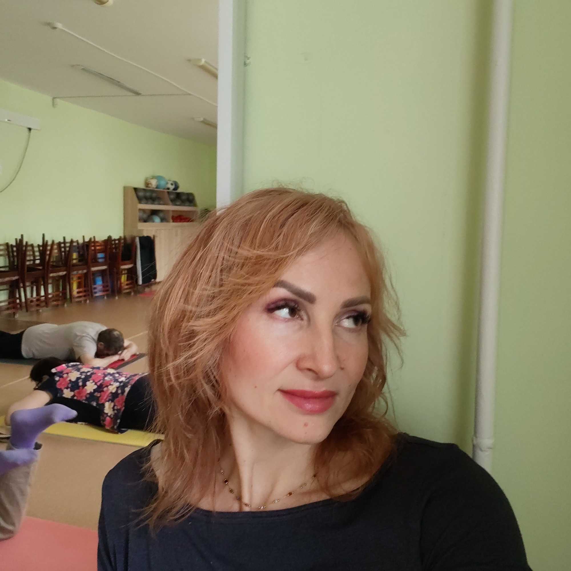 The image depicts a fair-skinned woman with wavy red-blonde hair, wearing a black top and a delicate beaded necklace. She has distinct eyebrows, eyelashes, and pink lip color. Her gaze is directed off to the side.
In the blurred background, several individuals are lying prone on mats. One person is wearing a gray shirt. Another person is wearing a floral shirt and purple socks. Chairs are stacked along the back wall of the room, adjacent to a shelf unit filled with items.
The room is painted a light green. The location is likely in a city with public buildings or centers where yoga is offered. It could be a dance studio.
Based on geolocation clues, this scene could be taking place in Chaikovsky, Perm Krai, Russia, where a yoga class or similar activity is occurring.